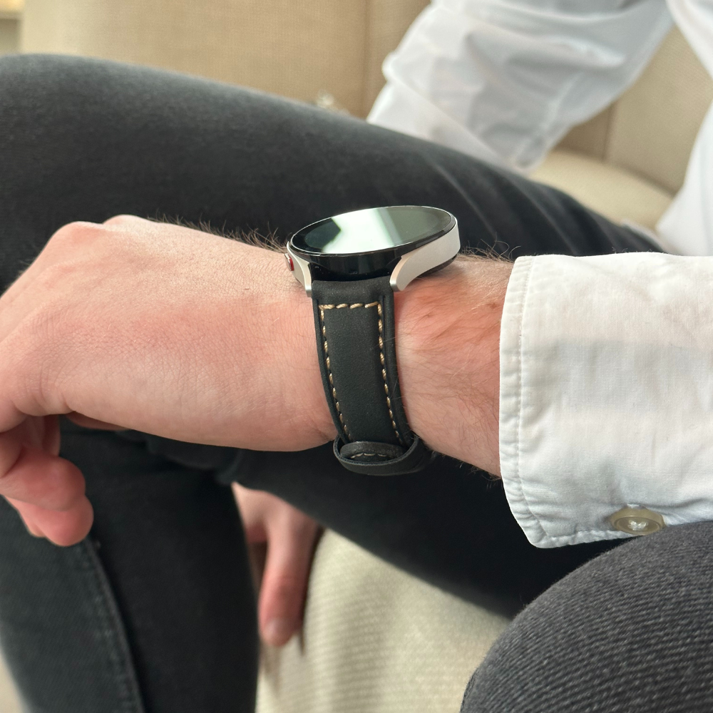 Das Handgelenk einer Person mit einer Smartwatch. Die Uhr hat ein schwarzes Lederarmband und ein silbernes und schwarzes Zifferblatt.