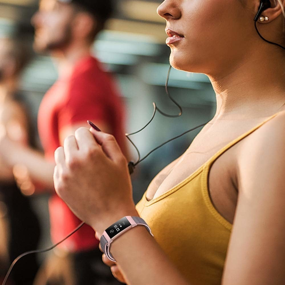 Frau in gelbem Oberteil mit Smartwatch, trägt Kopfhörer. Zwei Männer im Hintergrund. Sie hält das Kopfhörerkabel.