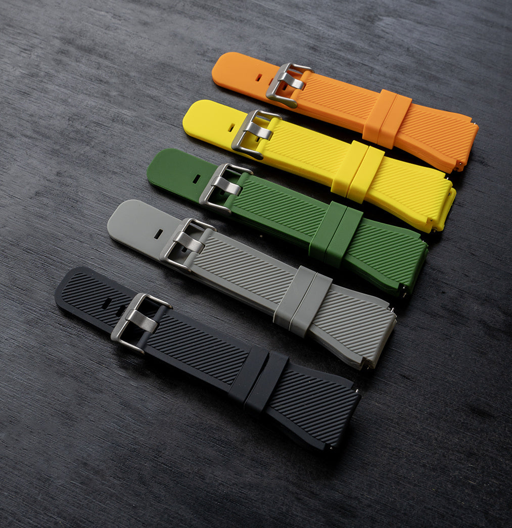 Fünf farbige Uhrenarmbänder diagonal auf dunkler Holzoberfläche angeordnet. Farben: Orange, Gelb, Grün, Grau und Schwarz.