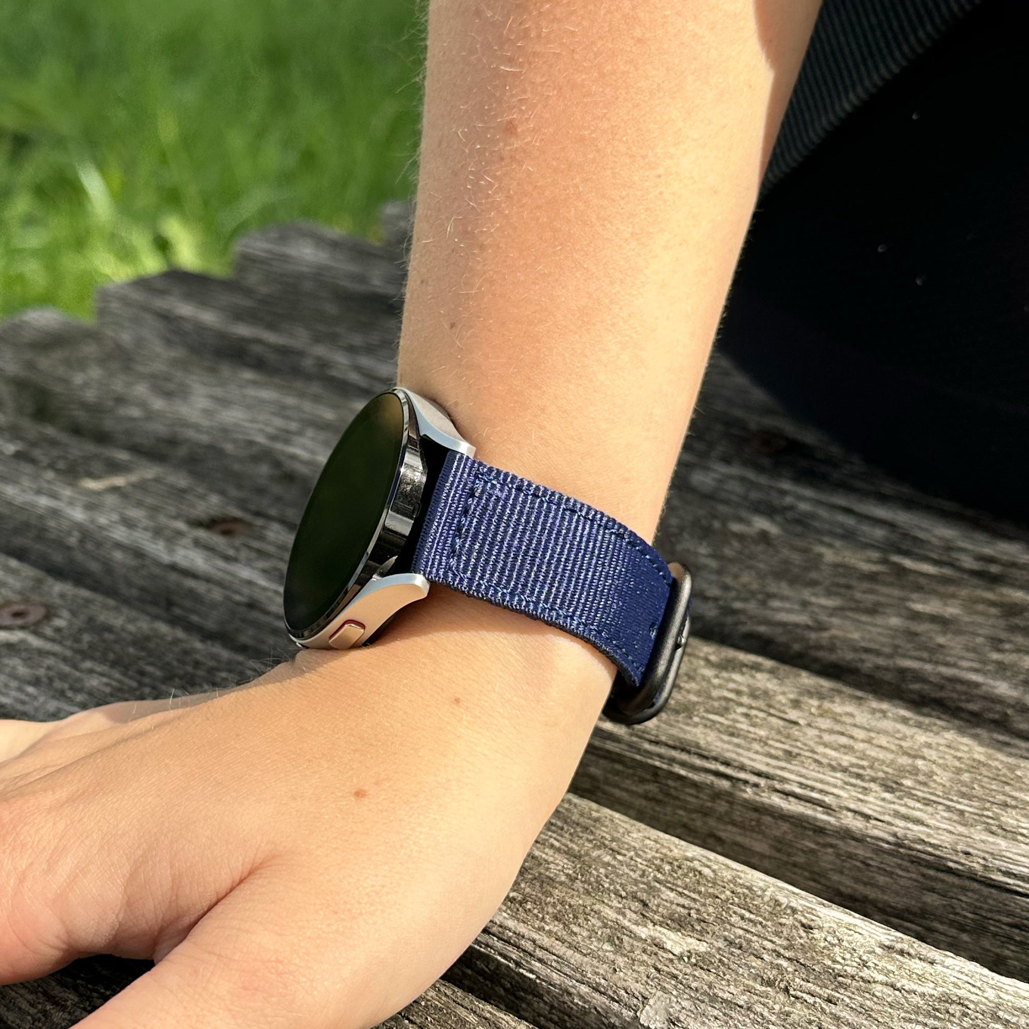 Ein Handgelenk mit einer silbernen Uhr und einem marineblauen Armband liegt auf einer Holzoberfläche. Grünes Gras ist im Hintergrund.