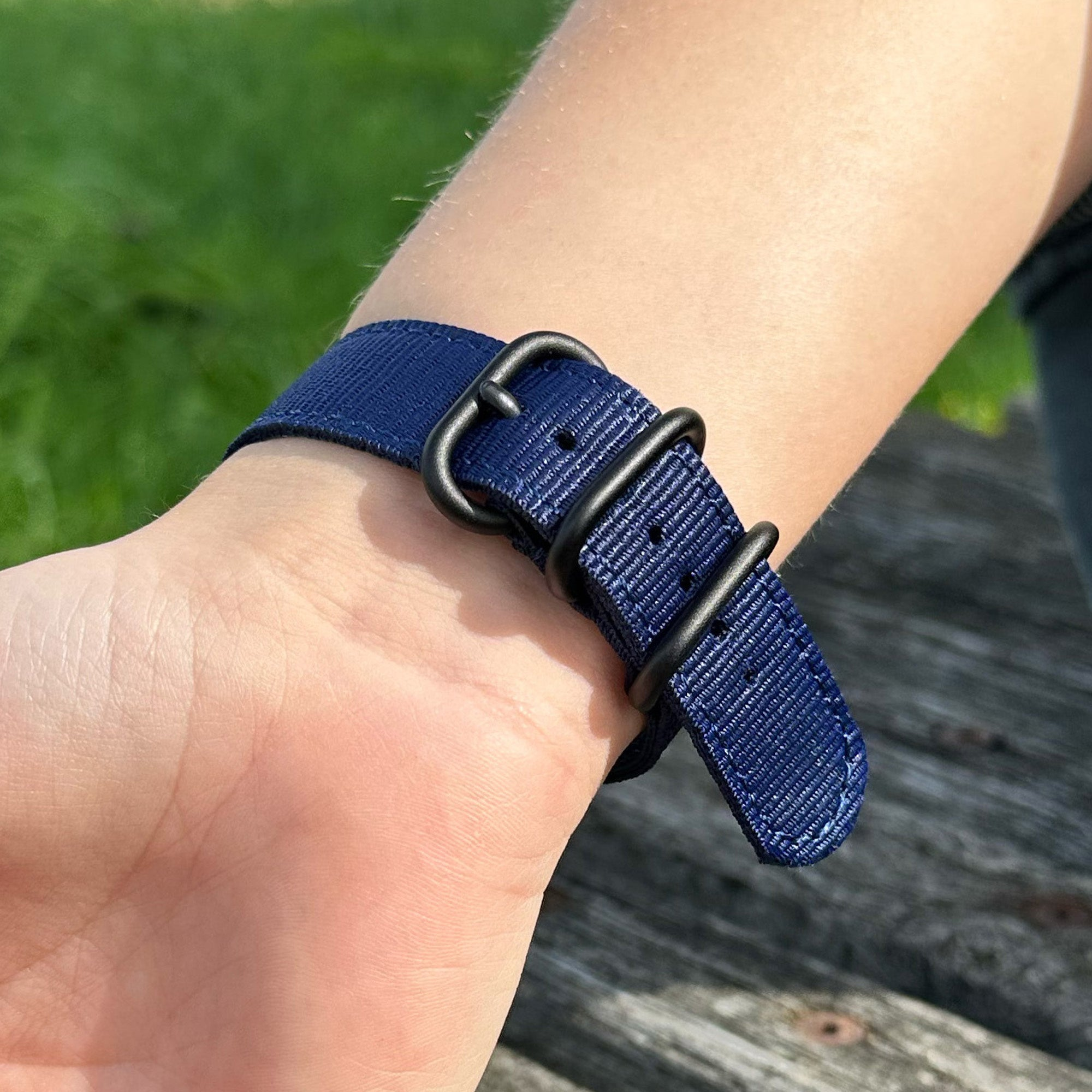 Nahaufnahme eines Handgelenks mit blauem Armband. Der schwarze Verschluss ist geschlossen. Grünes Gras und Holzplanken im Hintergrund.