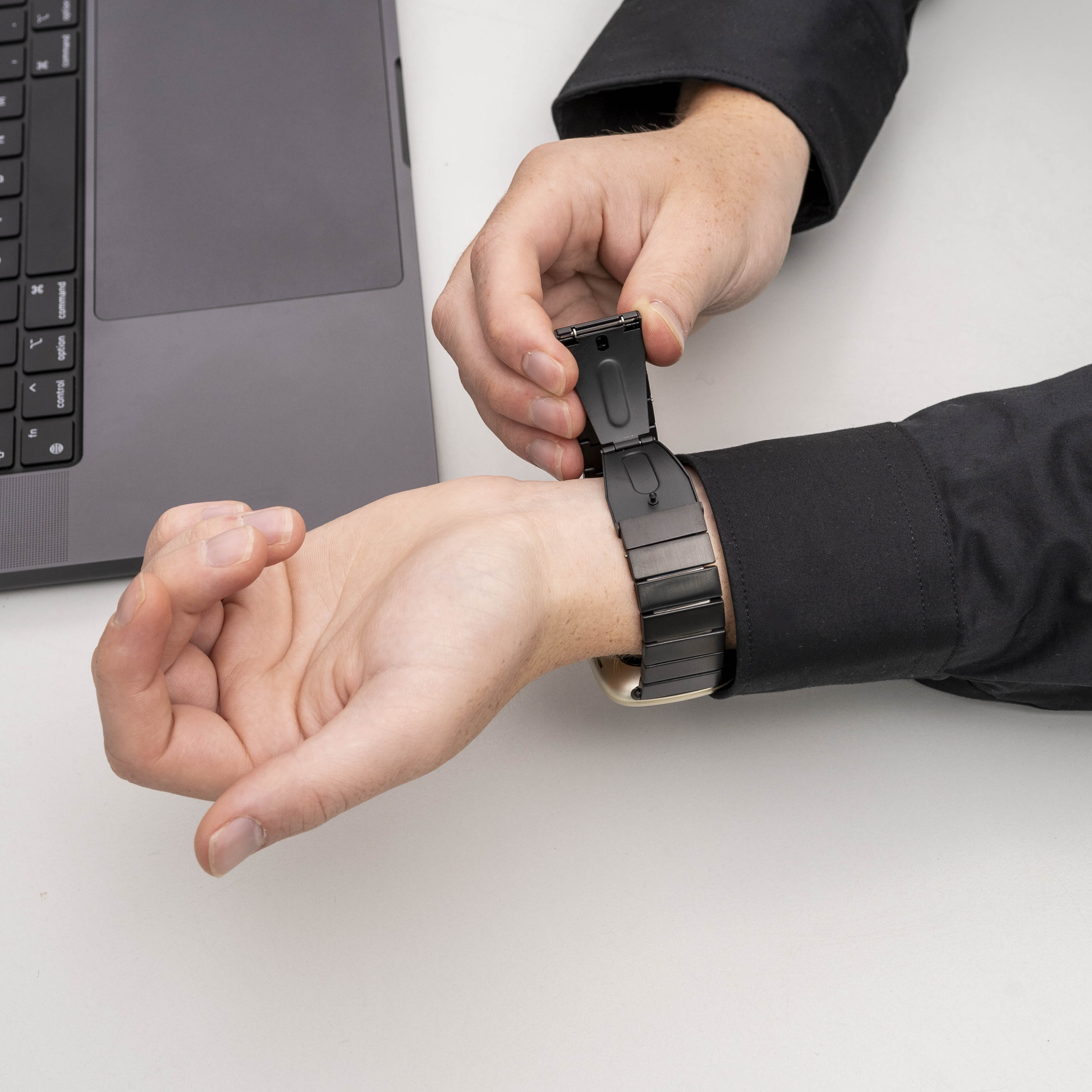 Eine Person befestigt eine schwarze Armbanduhr an ihrem Handgelenk. Links steht ein Laptop auf einer weißen Oberfläche.