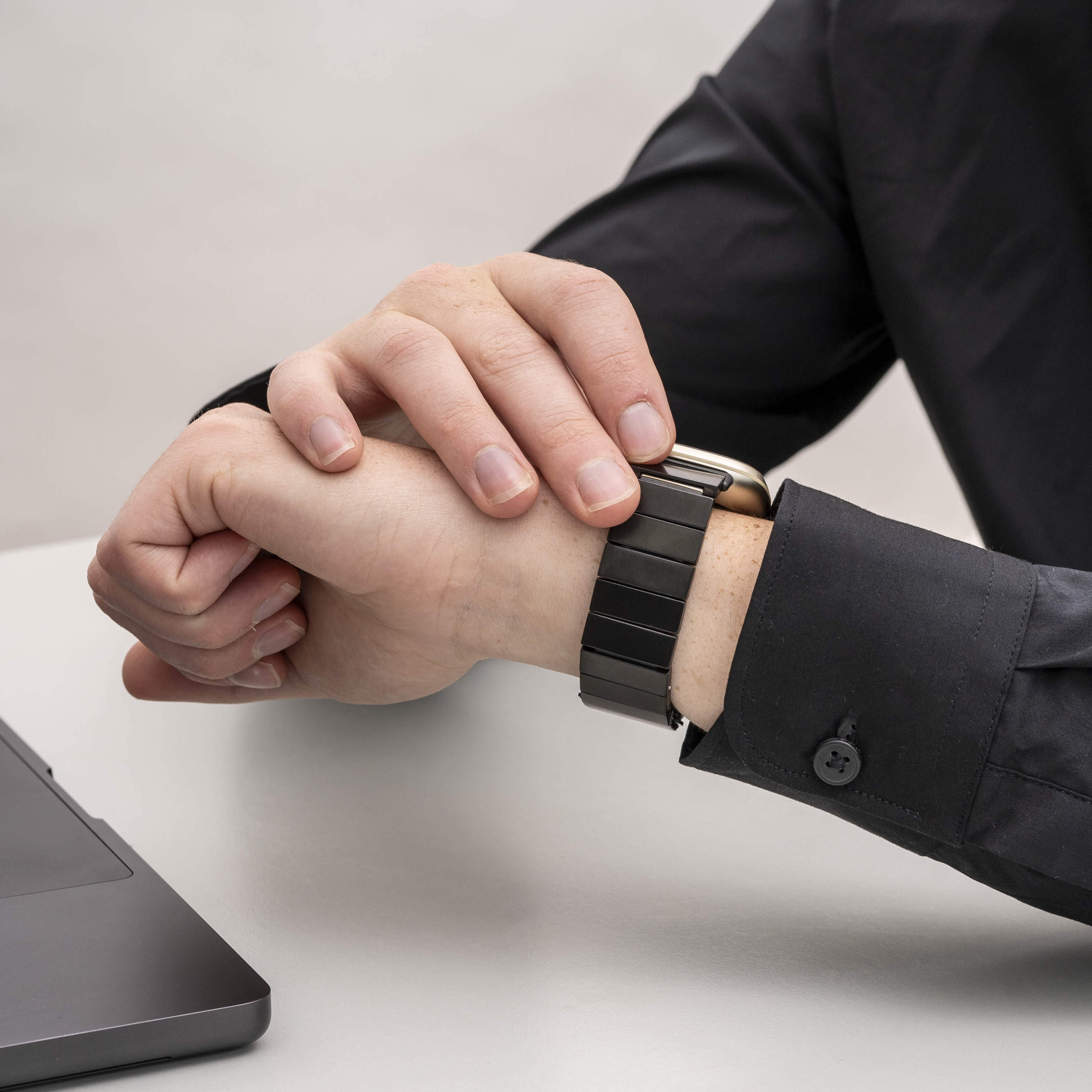 Handgelenk einer Person mit schwarzer Uhr und schwarzem Hemdärmel. Im Hintergrund ein Laptop.