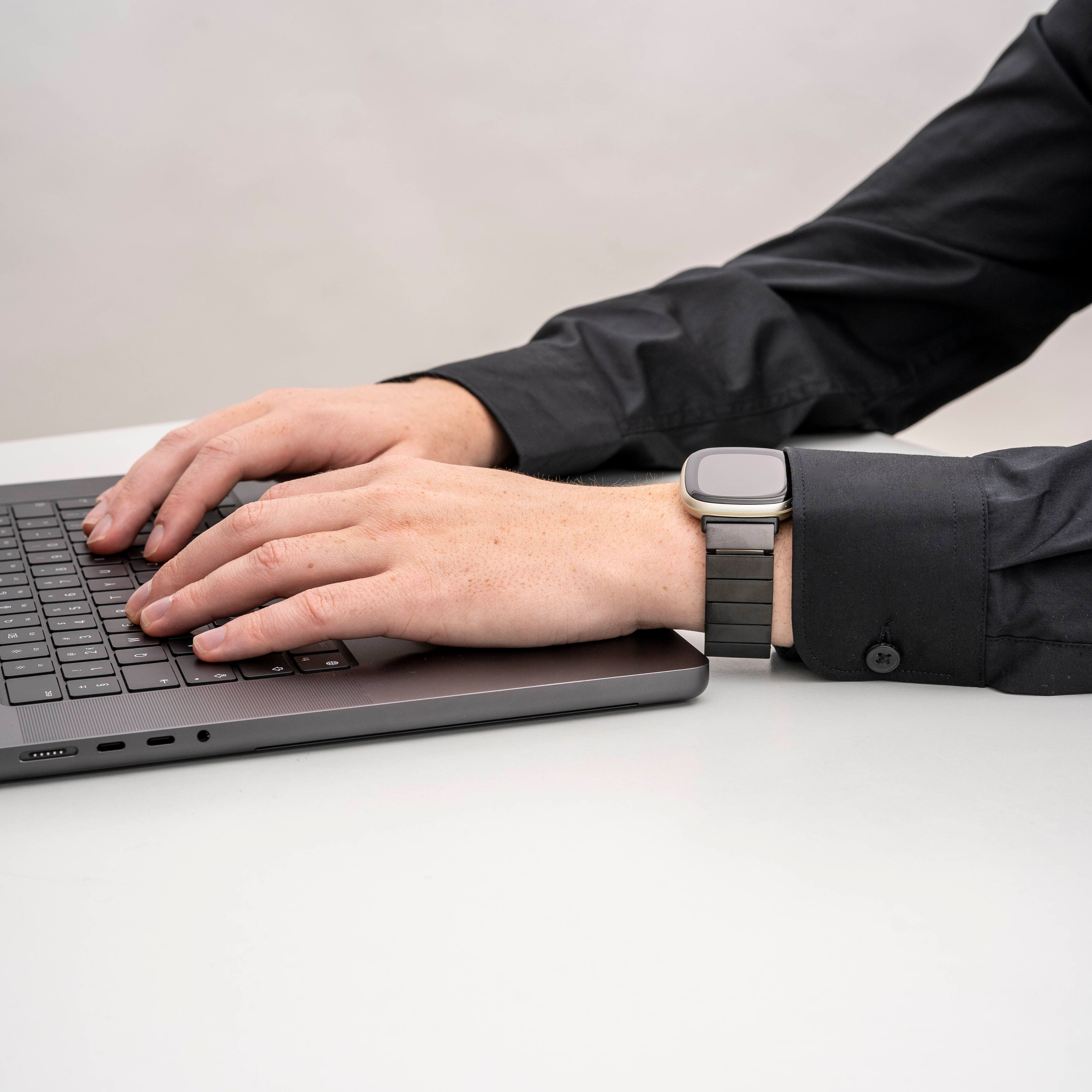 Eine Person tippt auf einem Laptop. Eine Smartwatch befindet sich an ihrem linken Handgelenk. Sie tragen ein schwarzes Hemd.