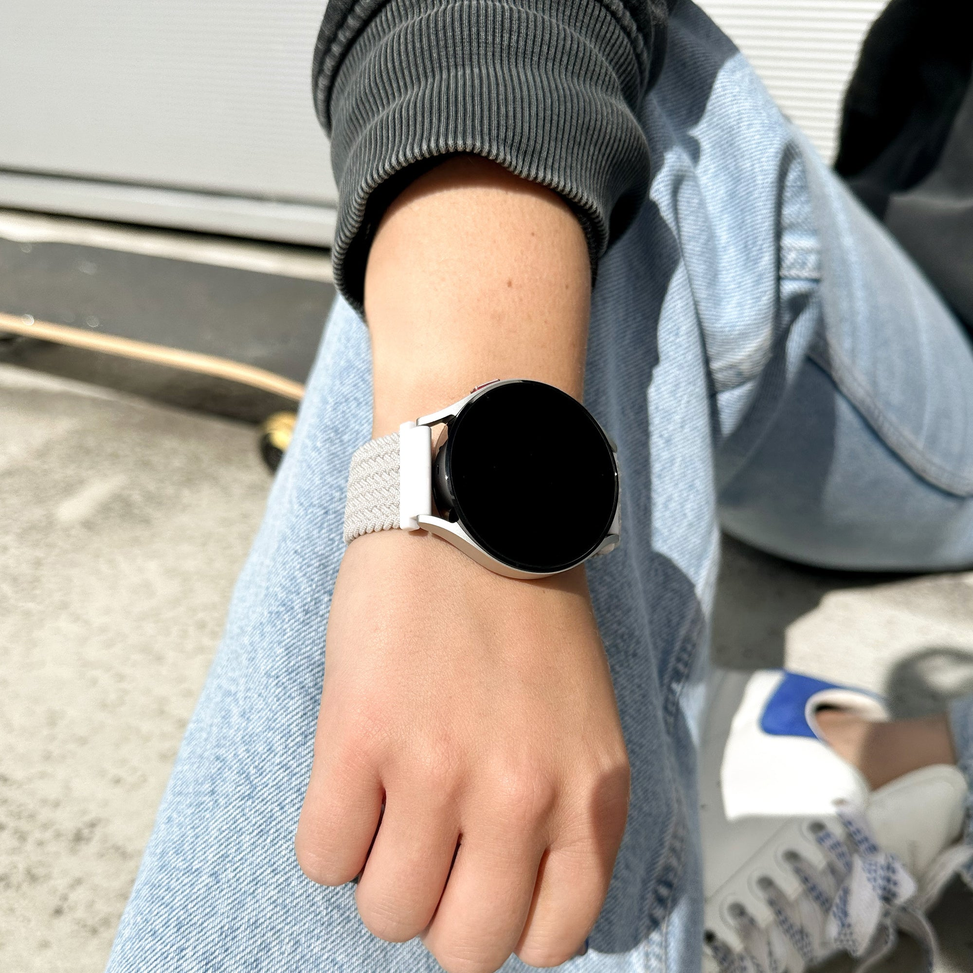 Person trägt Uhr, Jeans und Jacke. Im Hintergrund ein Skateboard und ein weißer Schuh mit blauen Akzenten.