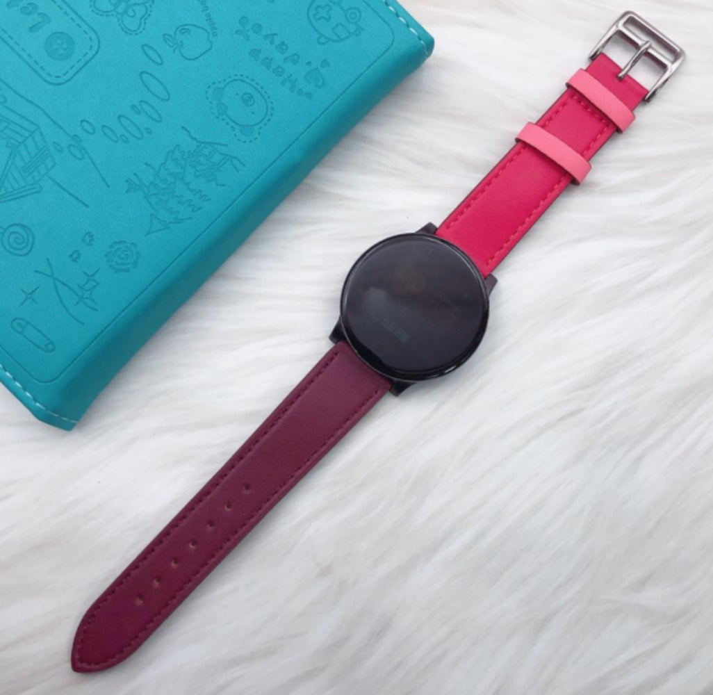 Eine schwarze Smartwatch mit rotem Armband liegt auf weißem Fell. Links liegt ein türkisfarbenes Notizbuch. Die Uhr hat ein rundes Zifferblatt.