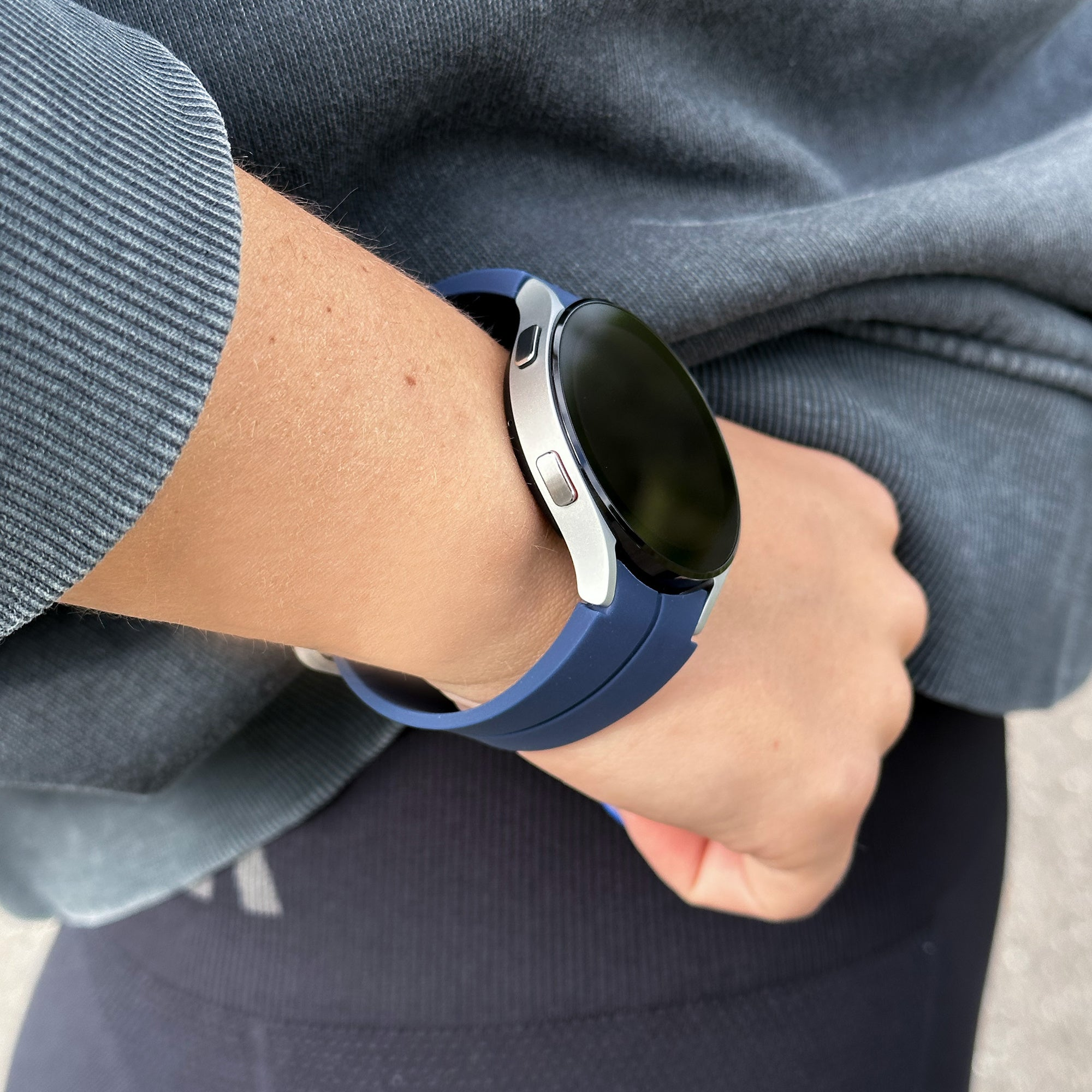 Nahaufnahme eines Handgelenks mit einer silbernen und blauen Smartwatch. Die Person trägt einen grauen Pullover und schwarze Leggings.