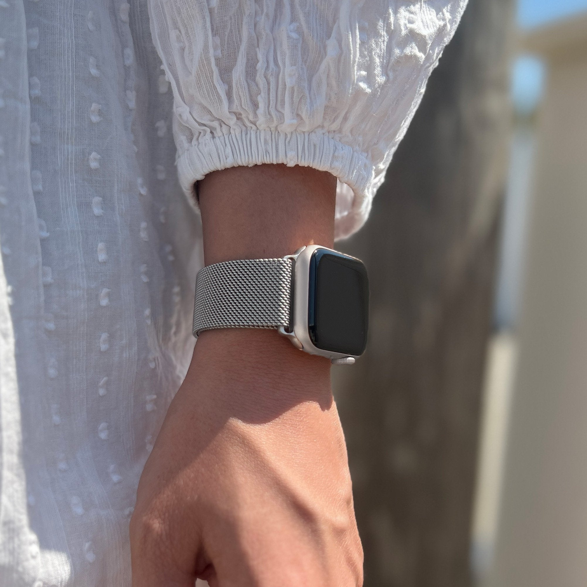 Arm einer Person mit silberner Uhr am Handgelenk. Sie trägt ein weißes Oberteil. Der Hintergrund ist unscharf.