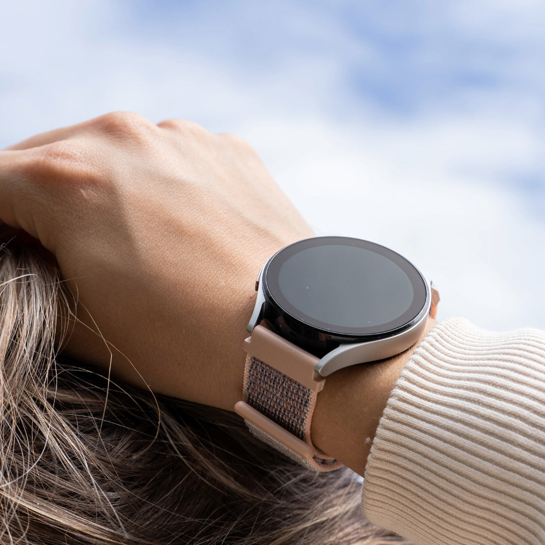 Handgelenk mit Smartwatch. Die Uhr hat einen schwarzen Bildschirm und ein rosa Armband. Der Hintergrund ist ein blauer Himmel.