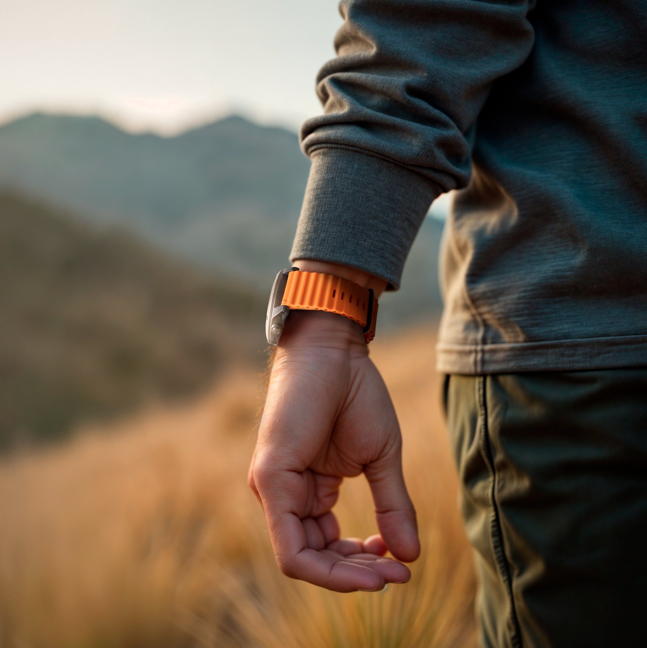 Ein Arm mit einer orangefarbenen Uhr, grüner Hose und einem grauen Hemd. Der Hintergrund ist ein verschwommenes Feld und Berge.