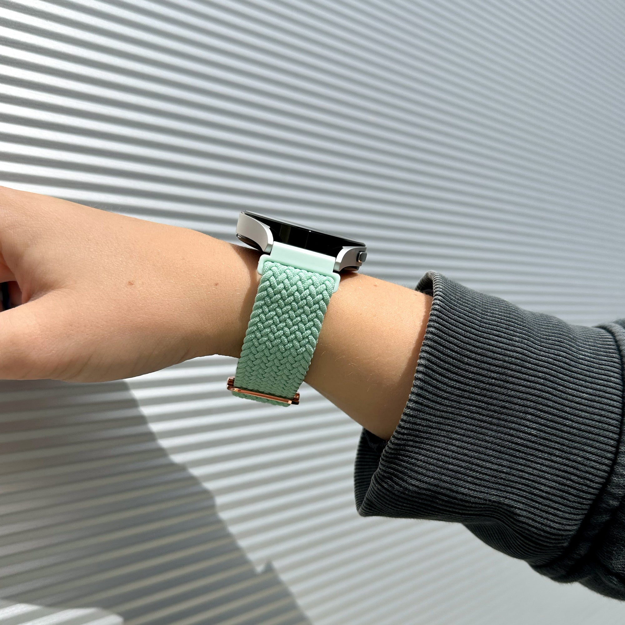 Das Handgelenk einer Person mit einem mintgrünen geflochtenen Uhrenarmband. Der Hintergrund ist eine metallische, geriffelte Wand.