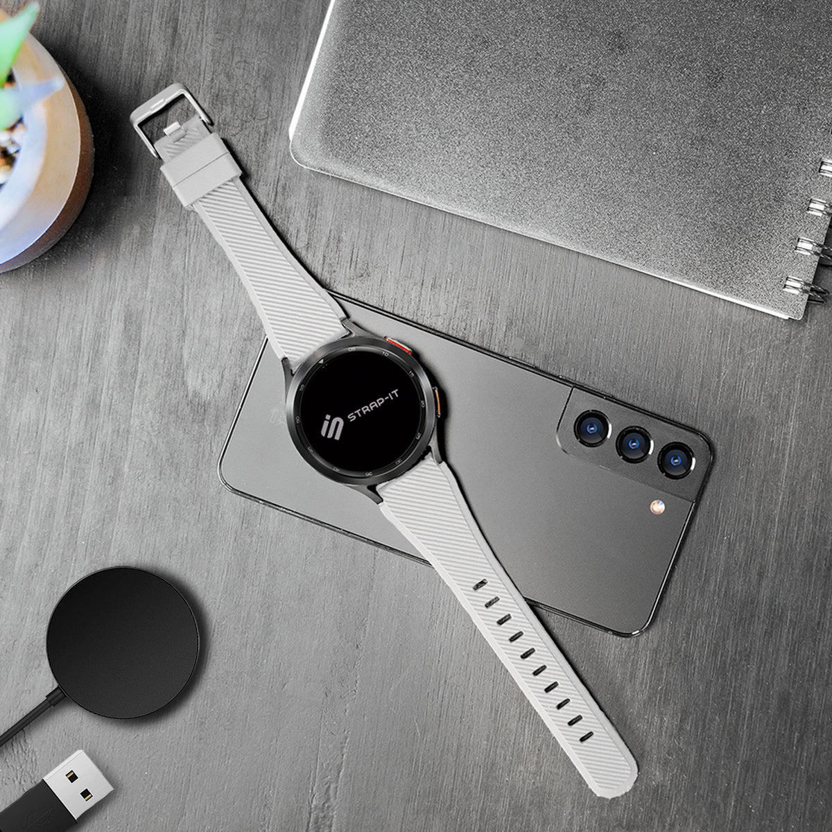 Eine Smartwatch mit hellgrauem Armband liegt auf einem grauen Smartphone. Links ist ein Ladegerät. Ein Notizbuch und eine Pflanze sind sichtbar.