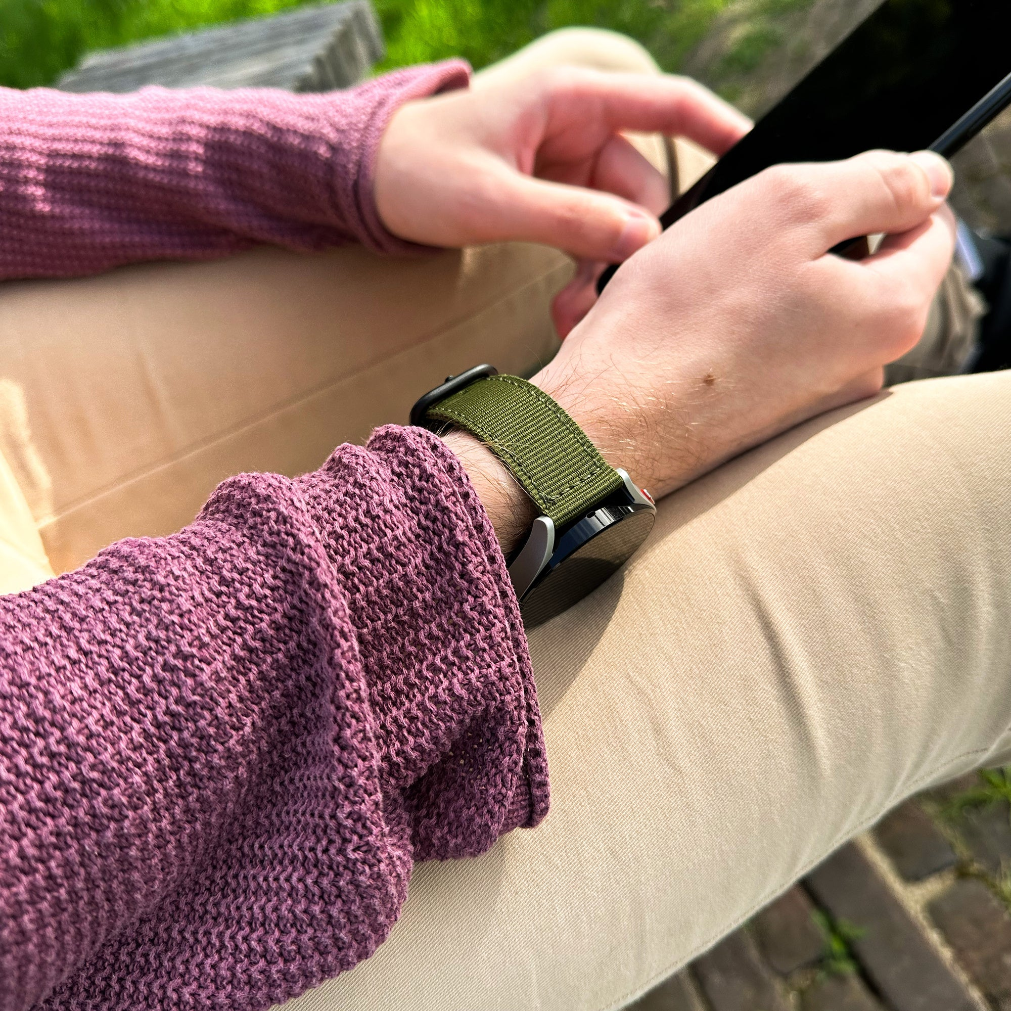 Person hält Telefon, trägt einen lila Pullover und eine grüne Uhr. Auch beige Hose und grüner Hintergrund sind sichtbar.