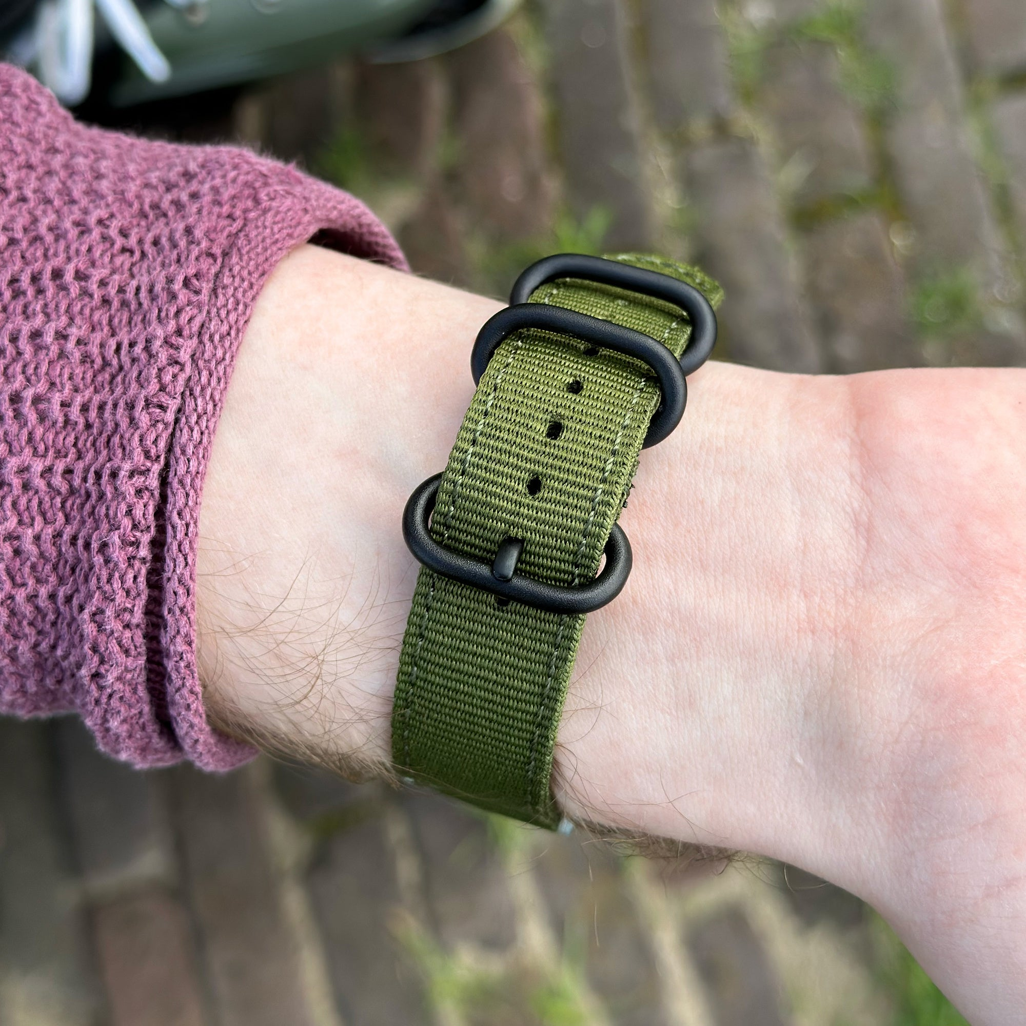 Ein Handgelenk mit einem grünen Uhrenarmband mit schwarzen Schnallen. Ein lila Ärmel ist sichtbar.