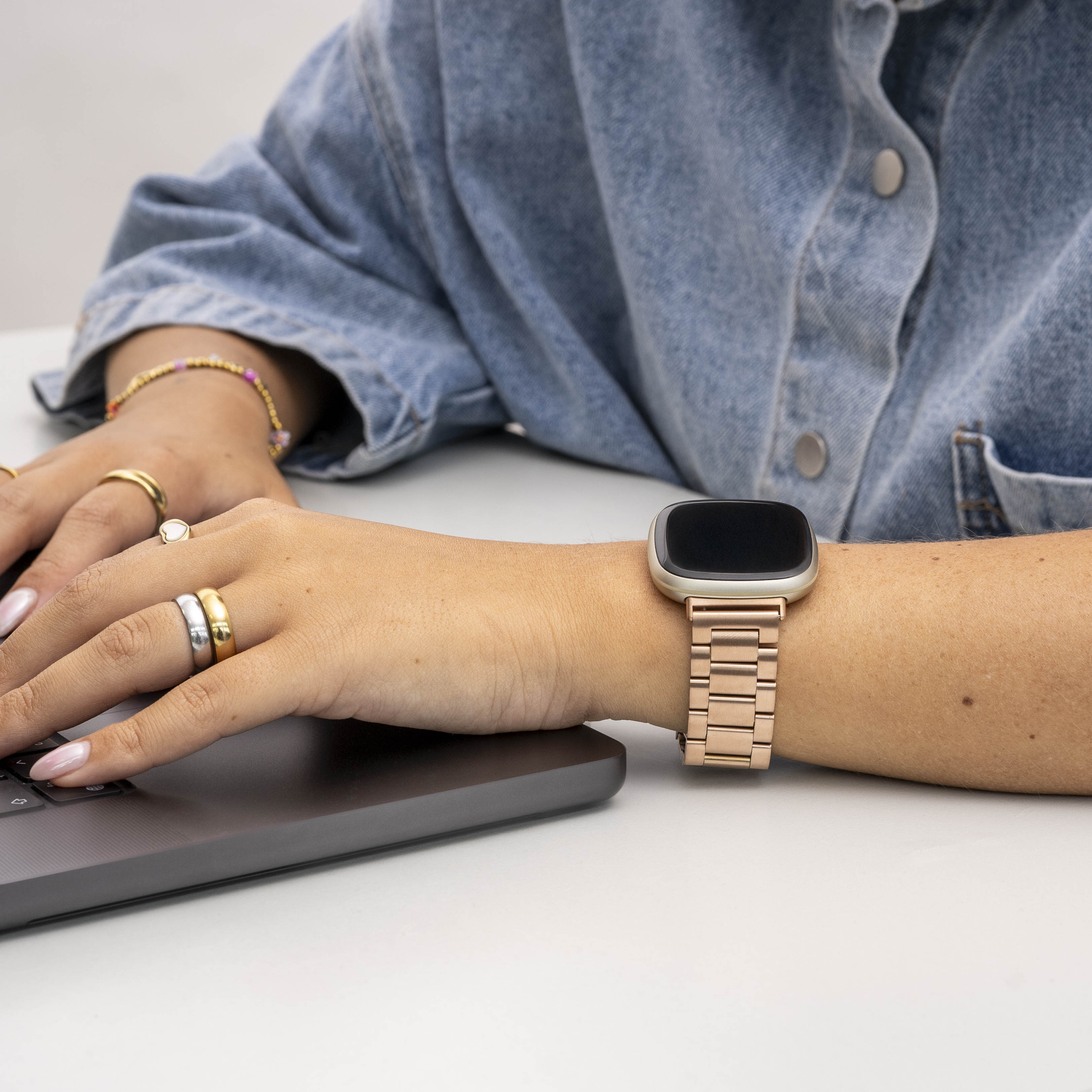 Person tippt auf einem Laptop. Sie trägt Ringe, eine roségoldene Uhr und eine Jeansjacke.