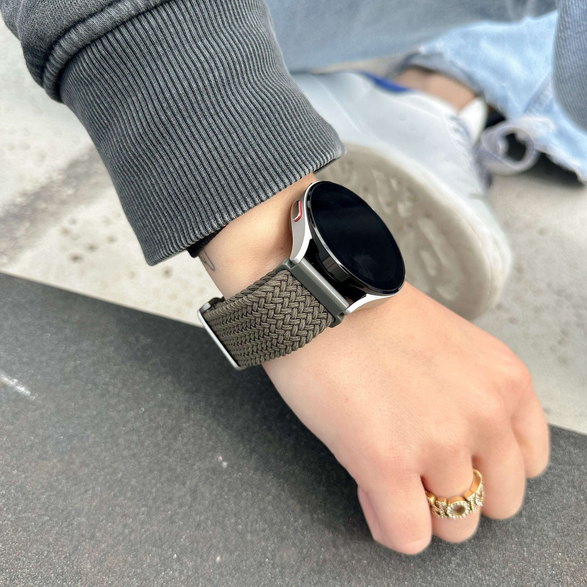 Eine Hand mit einer Uhr und einem Ring. Die Uhr hat ein schwarzes Zifferblatt und ein braunes Armband. Der Ring ist golden. Die Person trägt einen grauen Pullover.