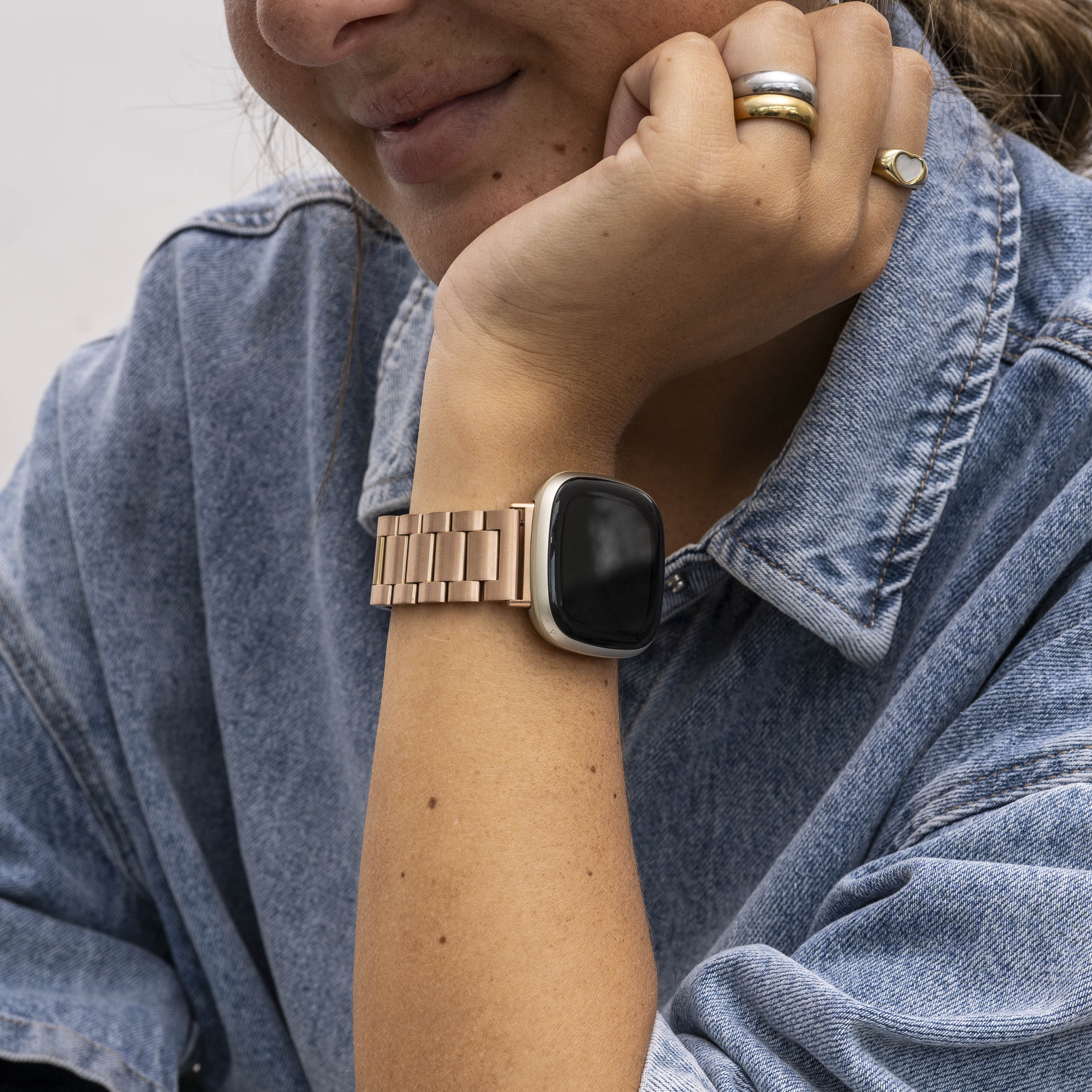 Der Arm einer Person mit einer roségoldenen Uhr. Sie trägt eine Jeansjacke und Ringe. Das Gesicht der Person ist ebenfalls sichtbar.