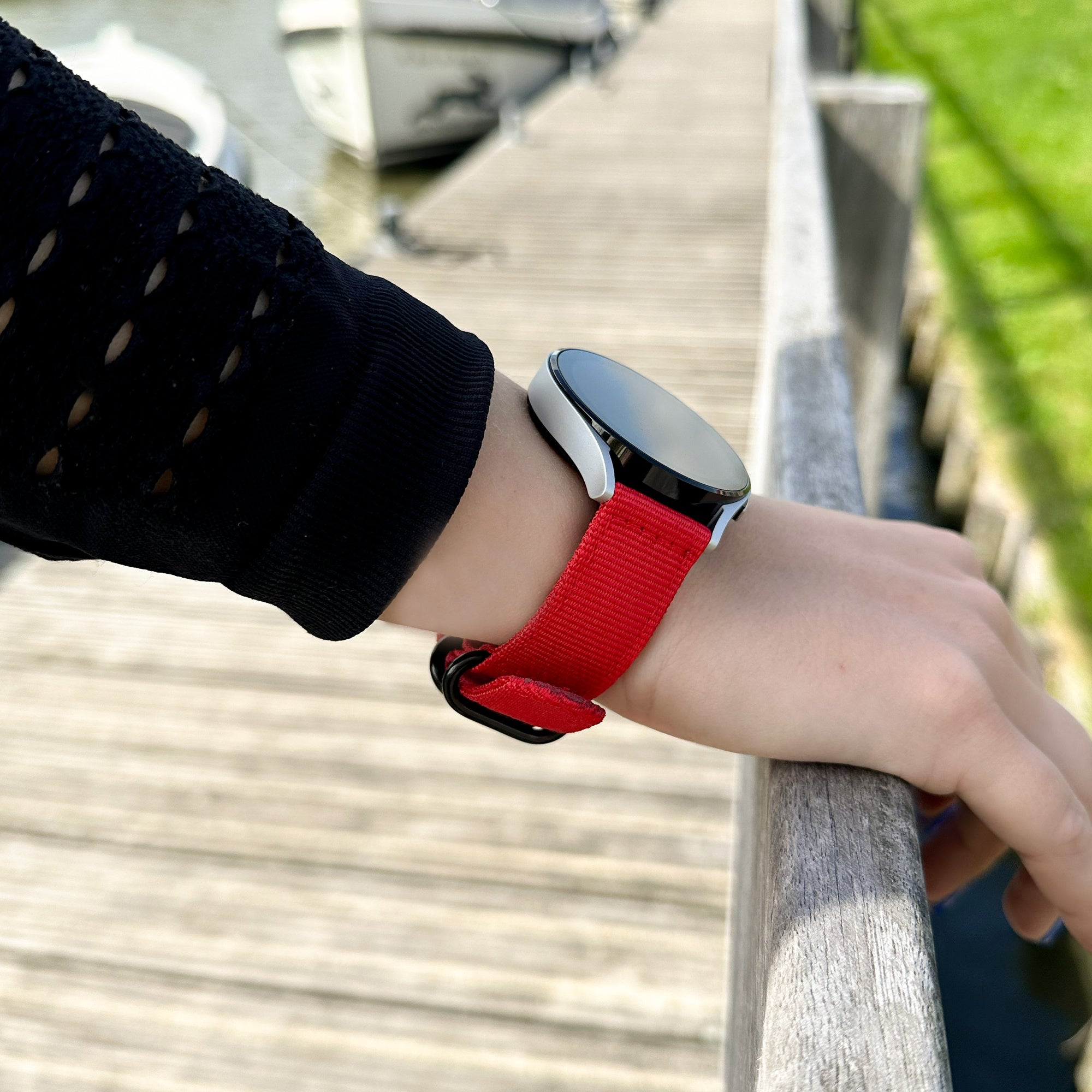 Das Handgelenk einer Person, die eine Smartwatch mit rotem Armband trägt, ruht auf einem Holzsteg. Boote im Hintergrund, grünes Gras.