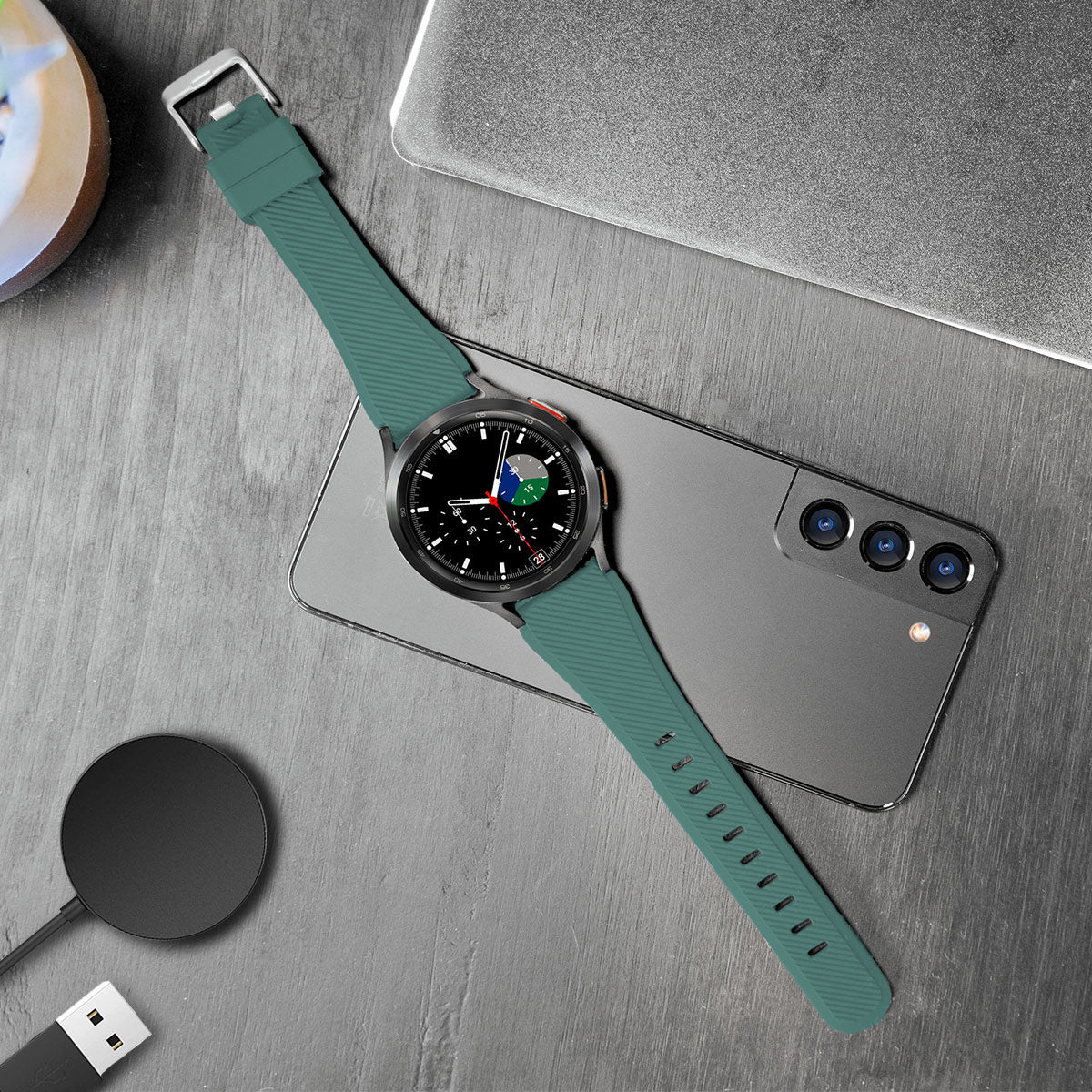 Eine tealfarbene Smartwatch mit schwarzem Zifferblatt liegt auf einem Telefon. Ein schwarzes kabelloses Ladegerät und ein Laptop sind im Hintergrund.