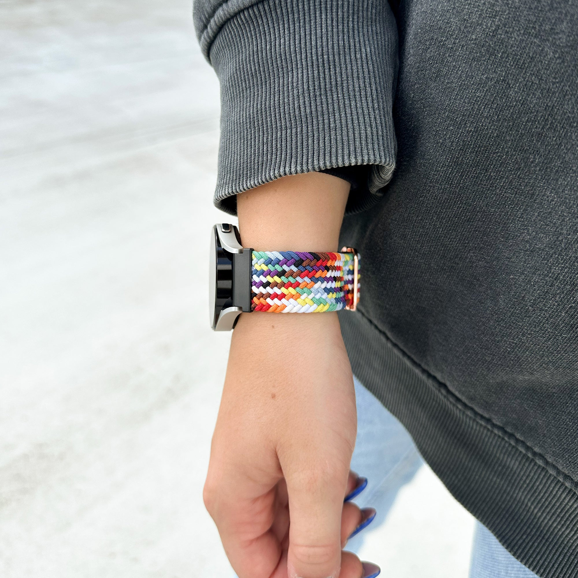 Ein Handgelenk mit einem farbenfrohen Regenbogen-Uhrenarmband. Die Person trägt einen grauen Pullover und hellblaue Jeans. Die Nägel sind blau.