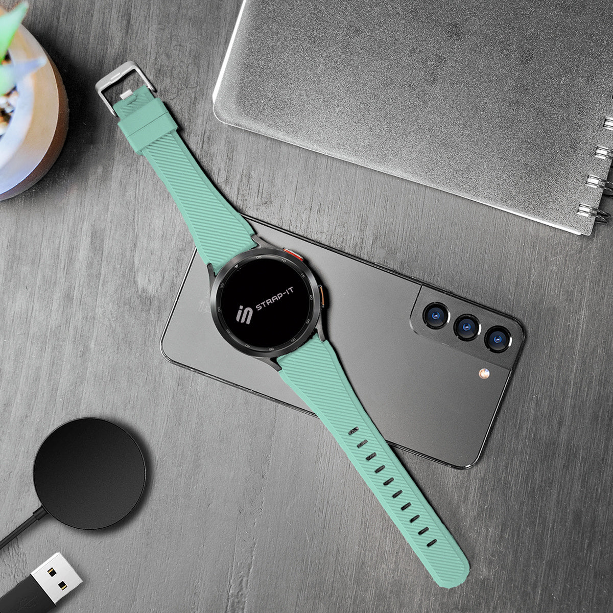 Eine Smartwatch mit tealfarbenem Armband liegt auf einem Telefon. Ein Ladegerät und ein Notizbuch sind ebenfalls auf einer grauen Oberfläche vorhanden.