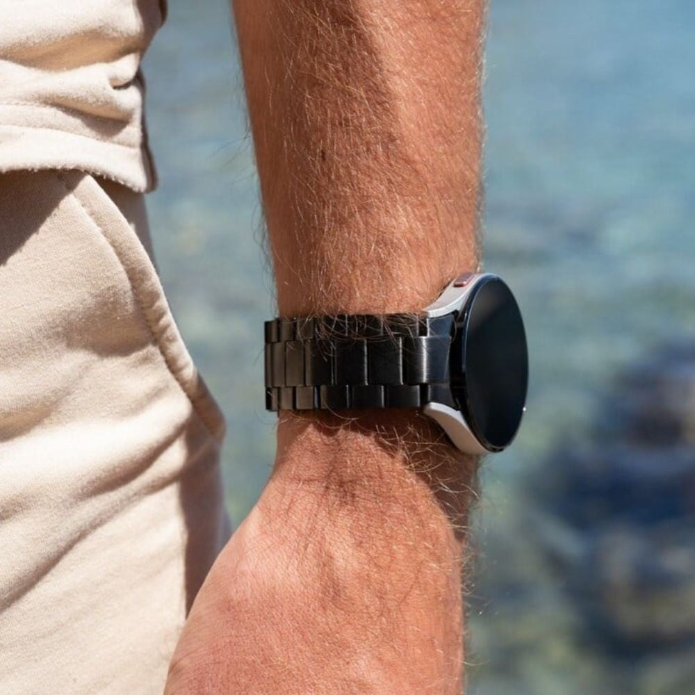 Ein Handgelenk mit einer schwarzen Uhr. Die Uhr hat eine silberne Lünette und ein schwarzes Armband. Der Hintergrund zeigt Wasser und Kleidung.