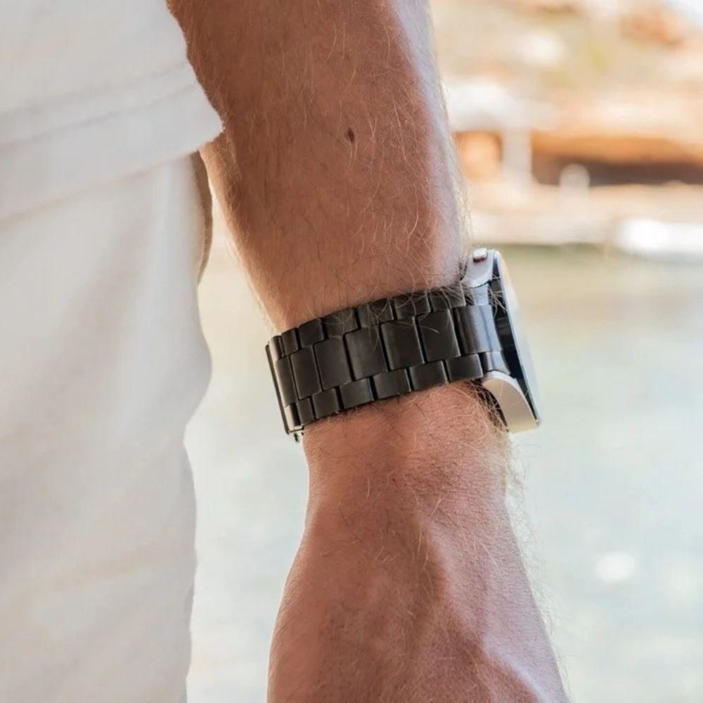 Der Arm eines Mannes mit einer schwarzen Metalluhr. Der Arm ist hellhäutig mit hellem Armhaar. Weißes Hemd sichtbar. Hintergrund ist verschwommen, im Freien.