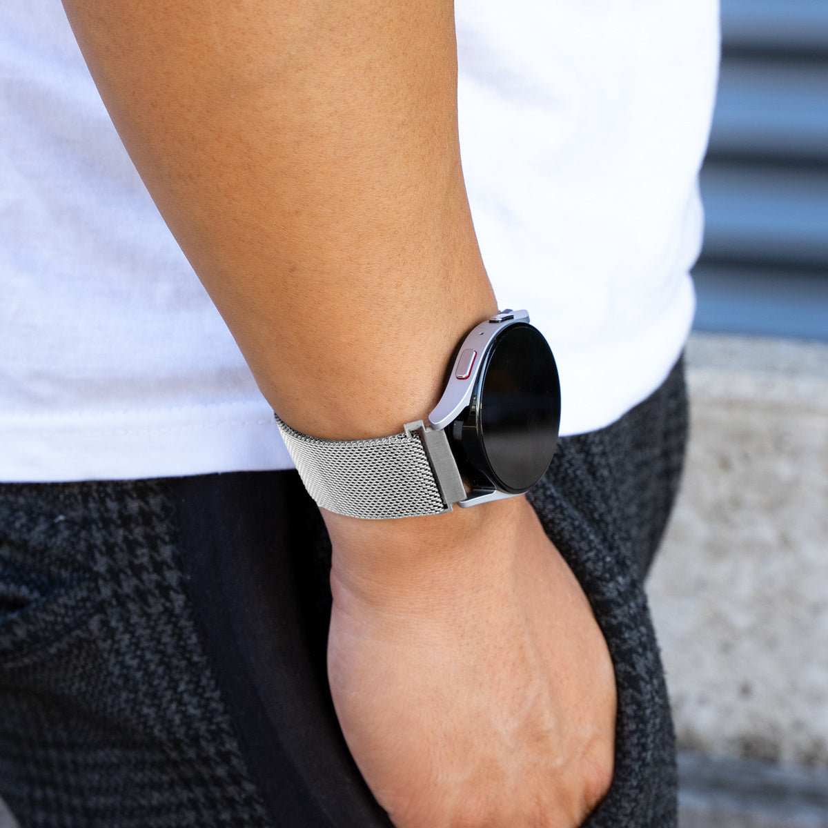 Person trägt eine silberne Smartwatch mit Mesh-Armband. Die Hand der Person ist in der Tasche. Ein weißes Hemd und eine graue Hose sind sichtbar.
