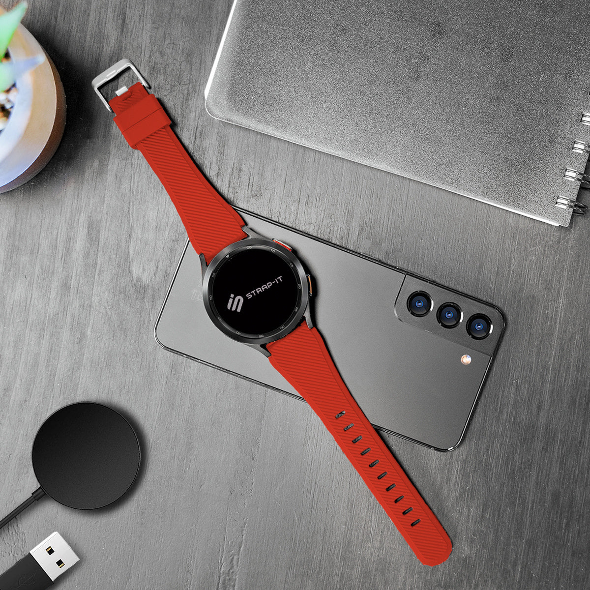 Eine Smartwatch mit rotem Armband liegt auf einem Telefon. Ein schwarzes Ladegerät, ein Notizbuch und eine Pflanze auf einem Holztisch.