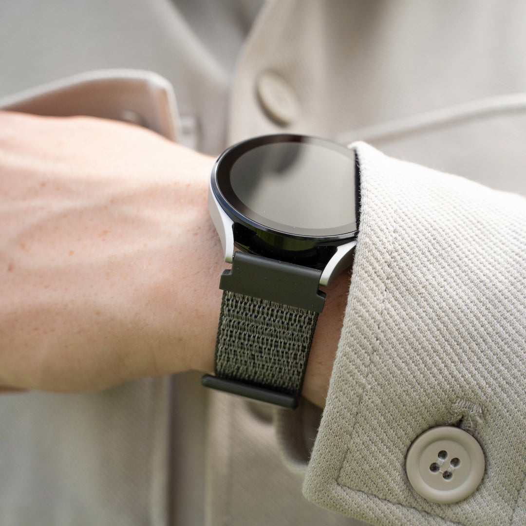 Eine Person trägt eine Smartwatch mit schwarzem Bildschirm und grünem Stoffband. Die Uhr befindet sich am Handgelenk, im Hintergrund eine beige Jacke.