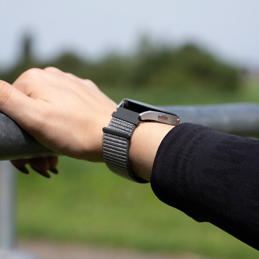 Handgelenk mit Smartwatch. Die Uhr hat ein graues Armband. Die Person trägt einen schwarzen Ärmel und hält eine graue Metallstange.