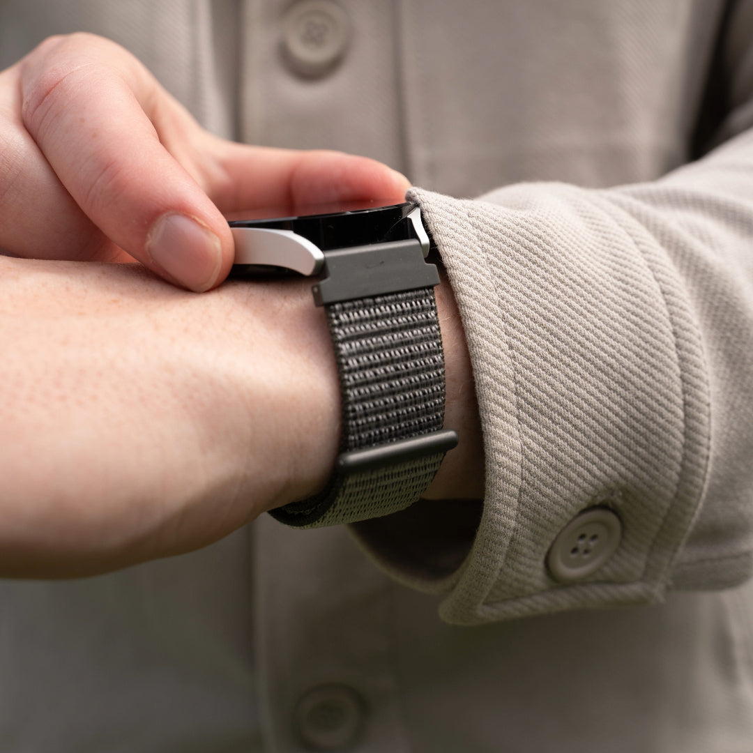 Eine Person justiert eine schwarze Uhr mit gewebtem Armband an ihrem Handgelenk. Sie trägt eine beige Jacke mit Knopf.