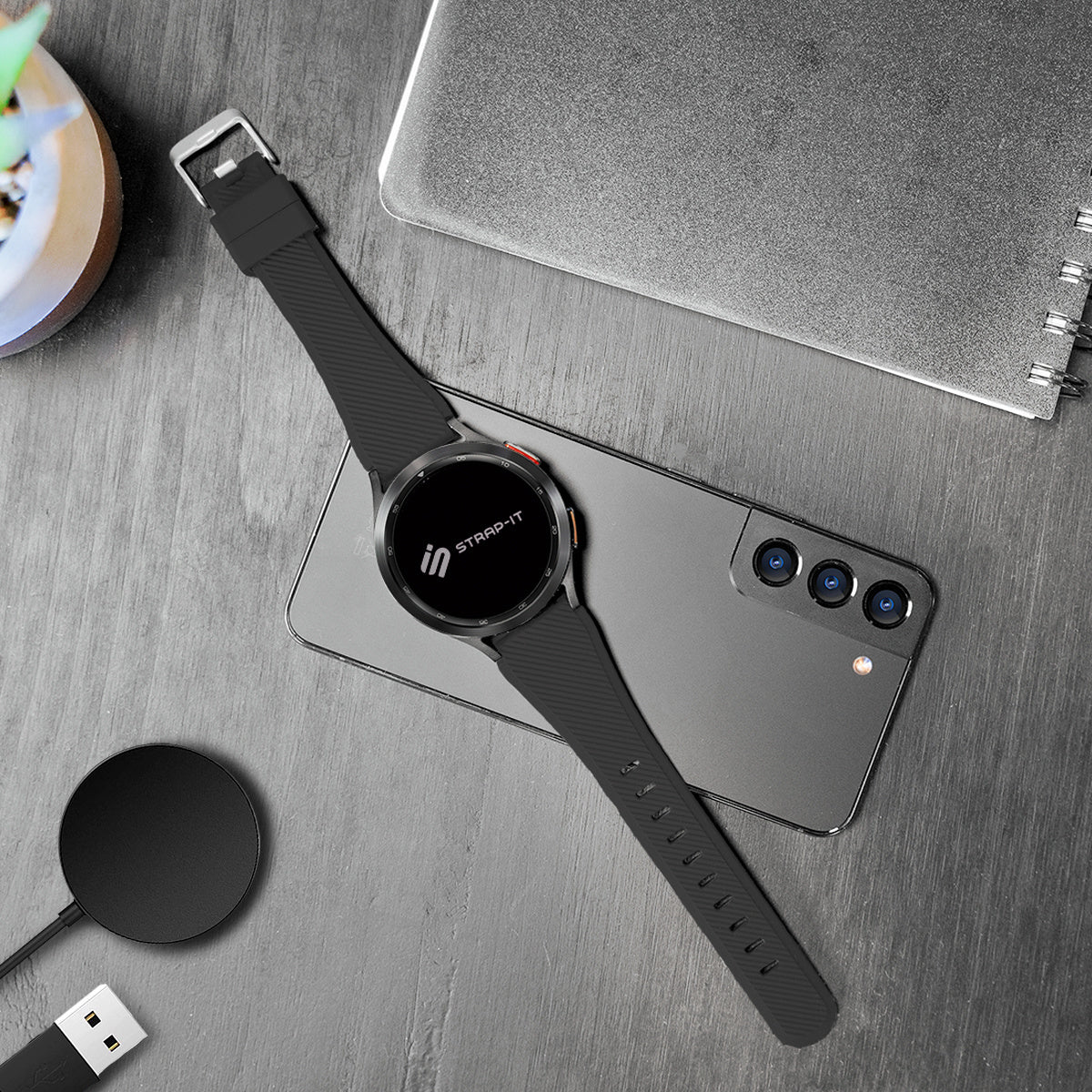 Eine schwarze Smartwatch mit schwarzem Armband liegt auf einem silbernen Telefon. Ein kabelloses Ladegerät und ein Notizbuch sind ebenfalls sichtbar.
