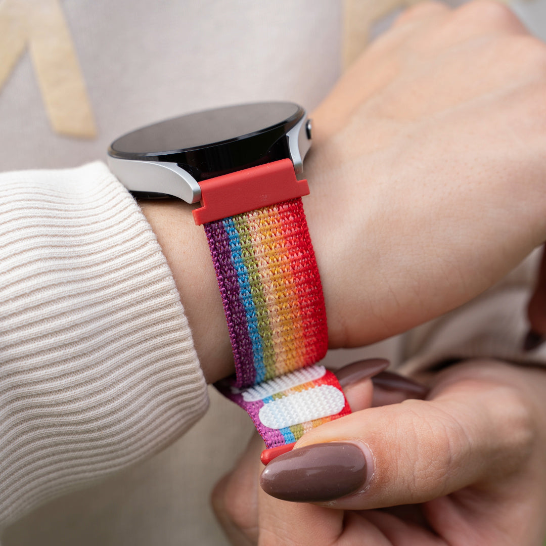 Eine Person trägt eine Smartwatch mit einem Regenbogenfarbenen Armband. Die Uhr ist silber und schwarz. Hände sind sichtbar.
