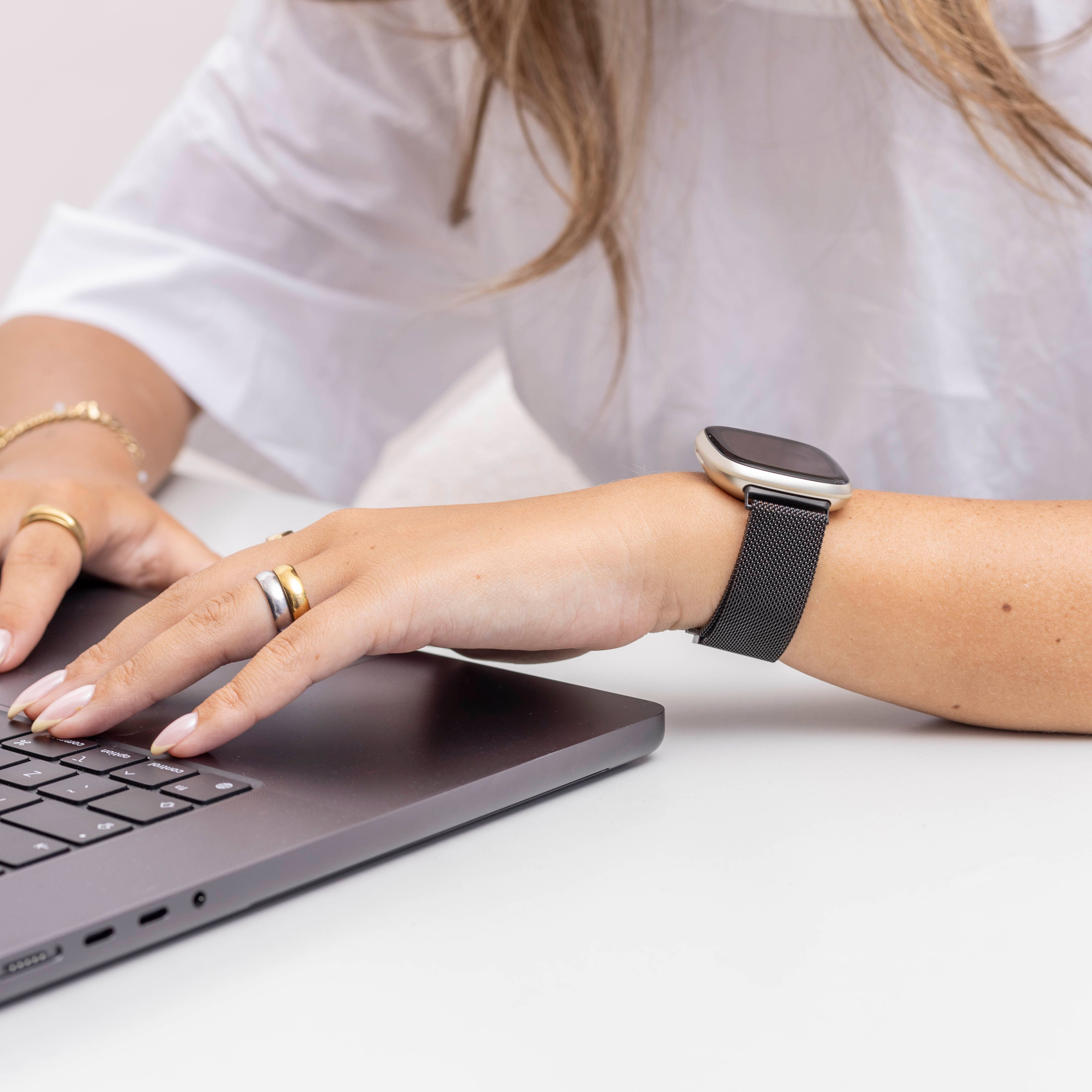 Eine Person tippt auf einem Laptop. Eine Smartwatch befindet sich am Handgelenk. Die Person trägt Ringe und ein weißes Hemd.