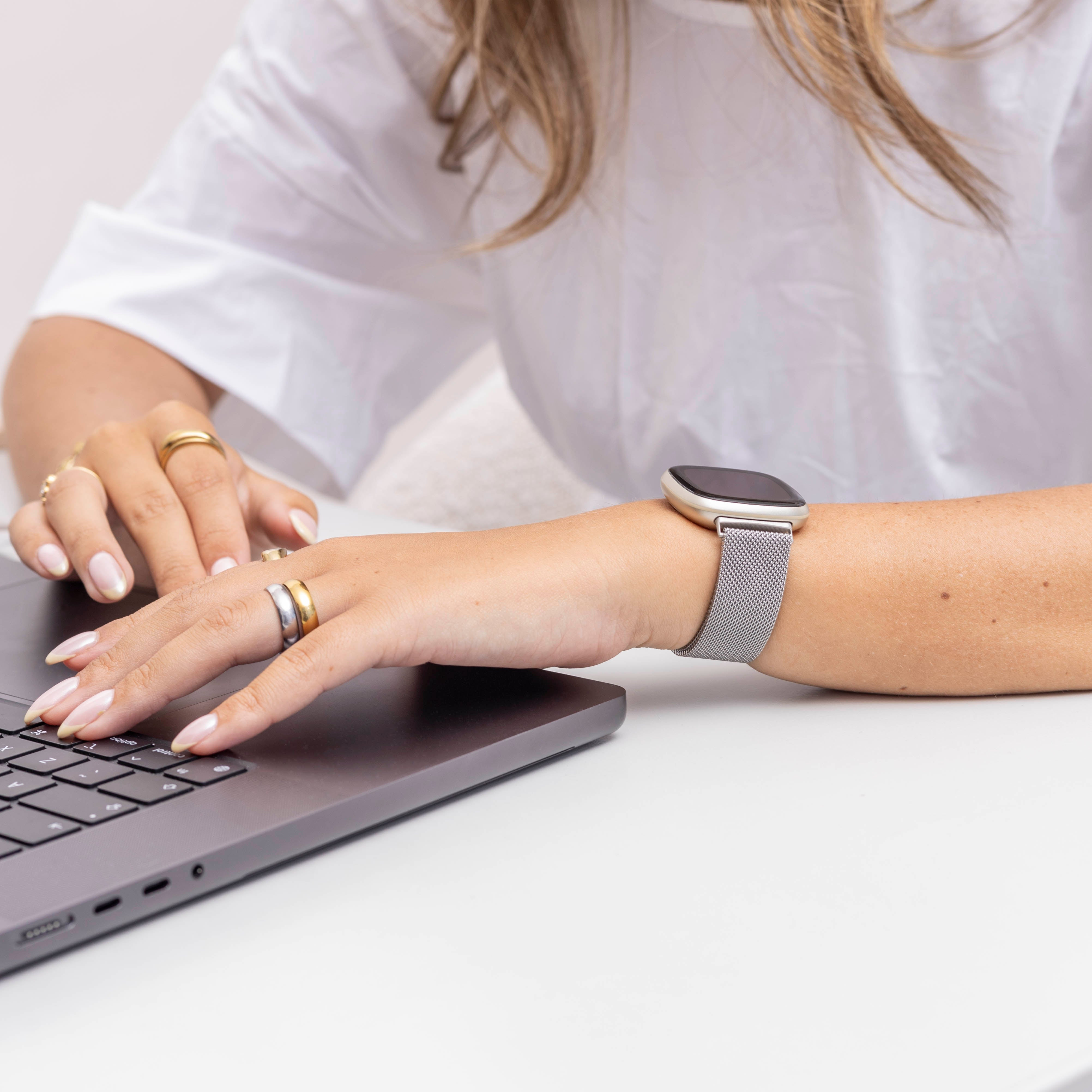 Hände einer Frau tippen auf einem Laptop. Sie trägt Ringe und eine Smartwatch. Ihre Nägel sind weiß lackiert.