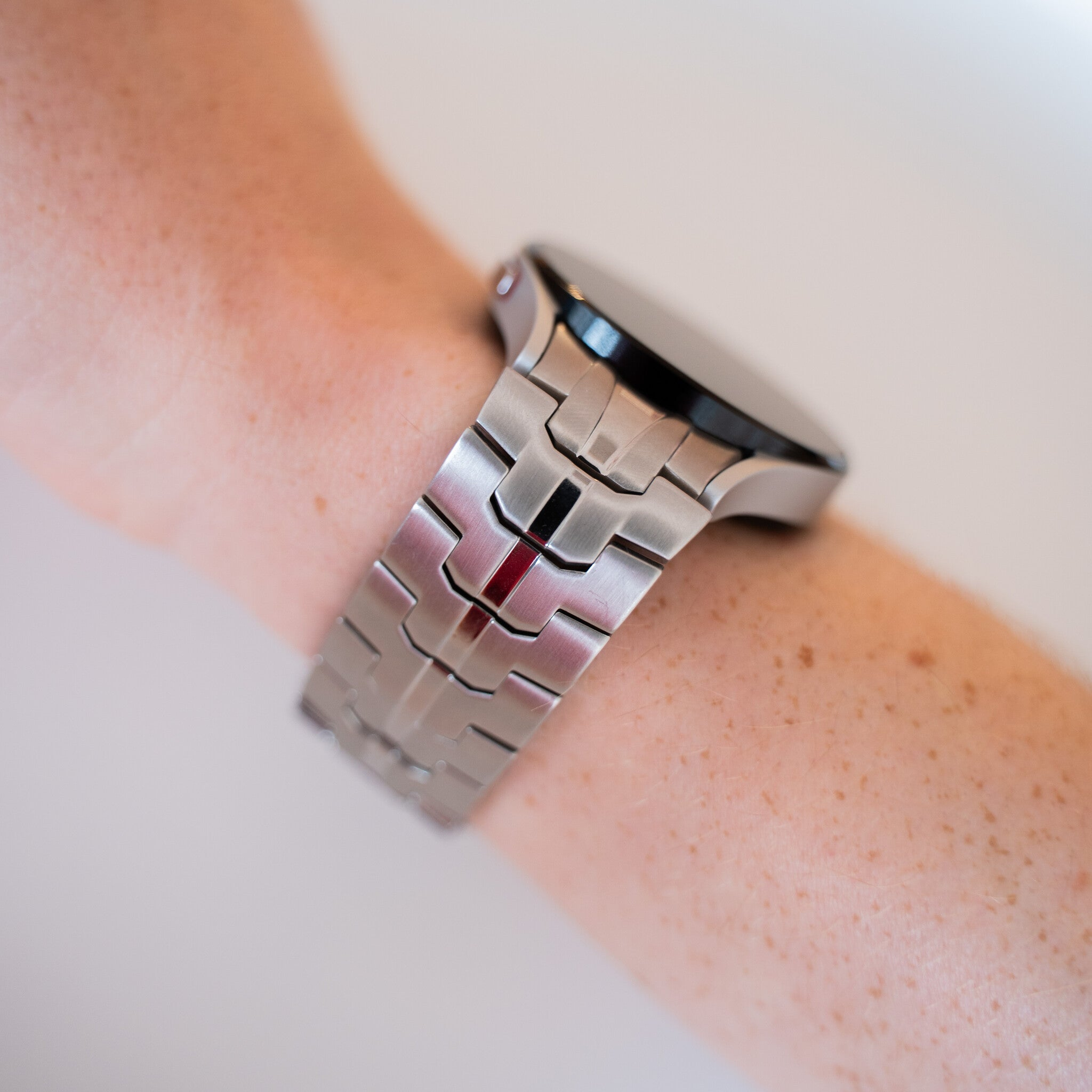 Eine silberne Smartwatch befindet sich an einem sommersprossigen Arm. Das Armband ist aus Metall. Der Hintergrund ist eine verschwommene weiße Oberfläche.