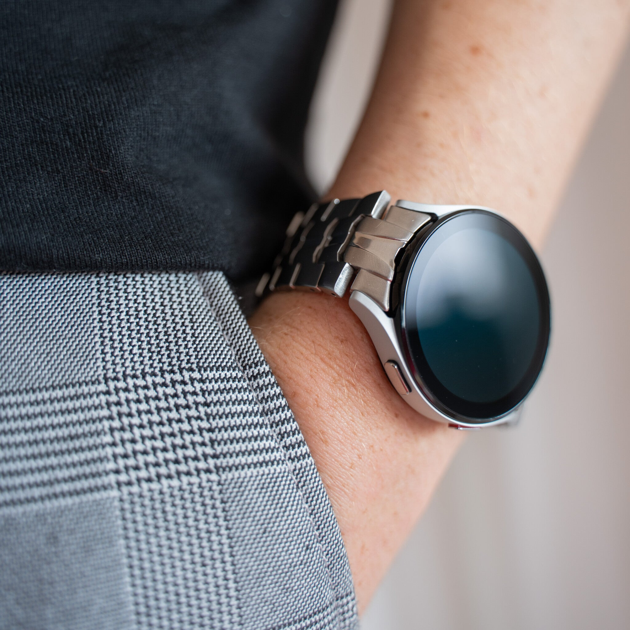 Das Handgelenk einer Person mit einer silbernen Smartwatch. Die Uhr hat ein schwarzes Zifferblatt und das Handgelenk befindet sich neben einem grauen Karostoff.