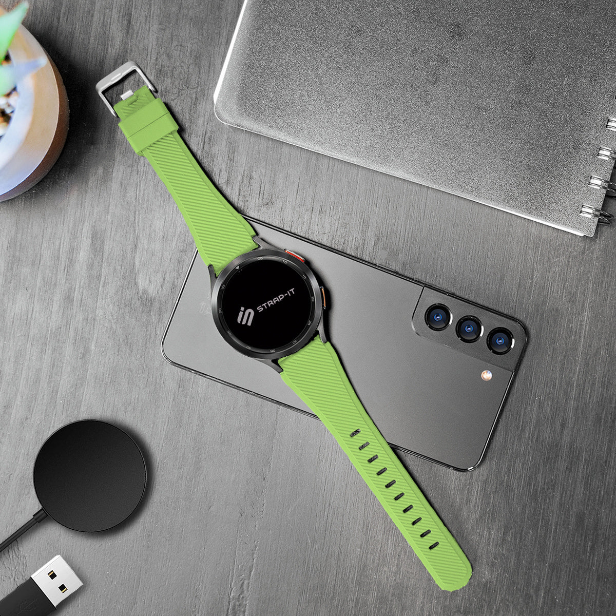 Eine Smartwatch mit grünem Armband liegt auf einem Smartphone. Weitere Gegenstände sind ein Ladepad, ein Notizbuch und eine kleine Pflanze.