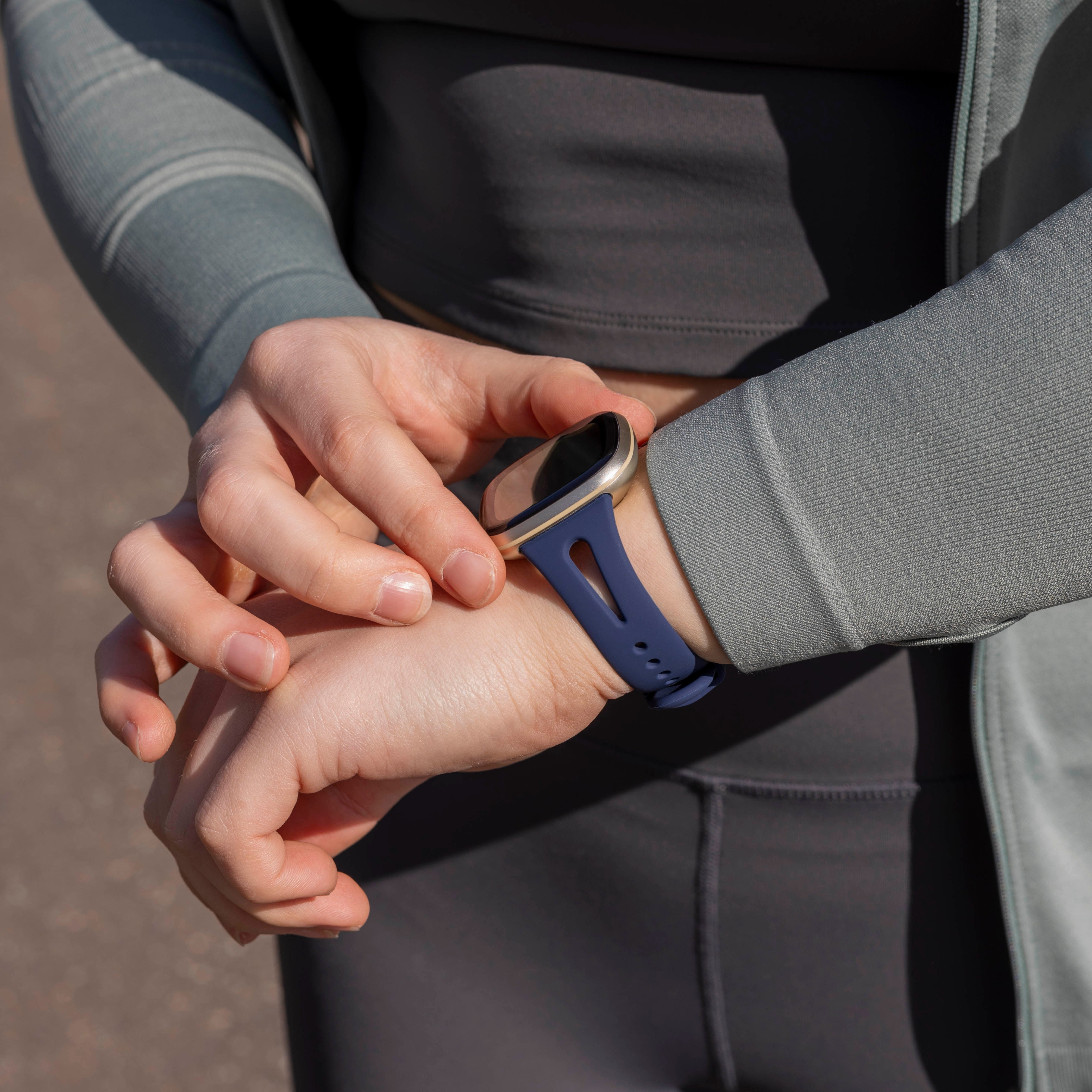 Person, die eine Smartwatch trägt. Hände überprüfen die Uhr mit blauem Armband. Dunkle Kleidung und eine graue Jacke sind ebenfalls sichtbar.