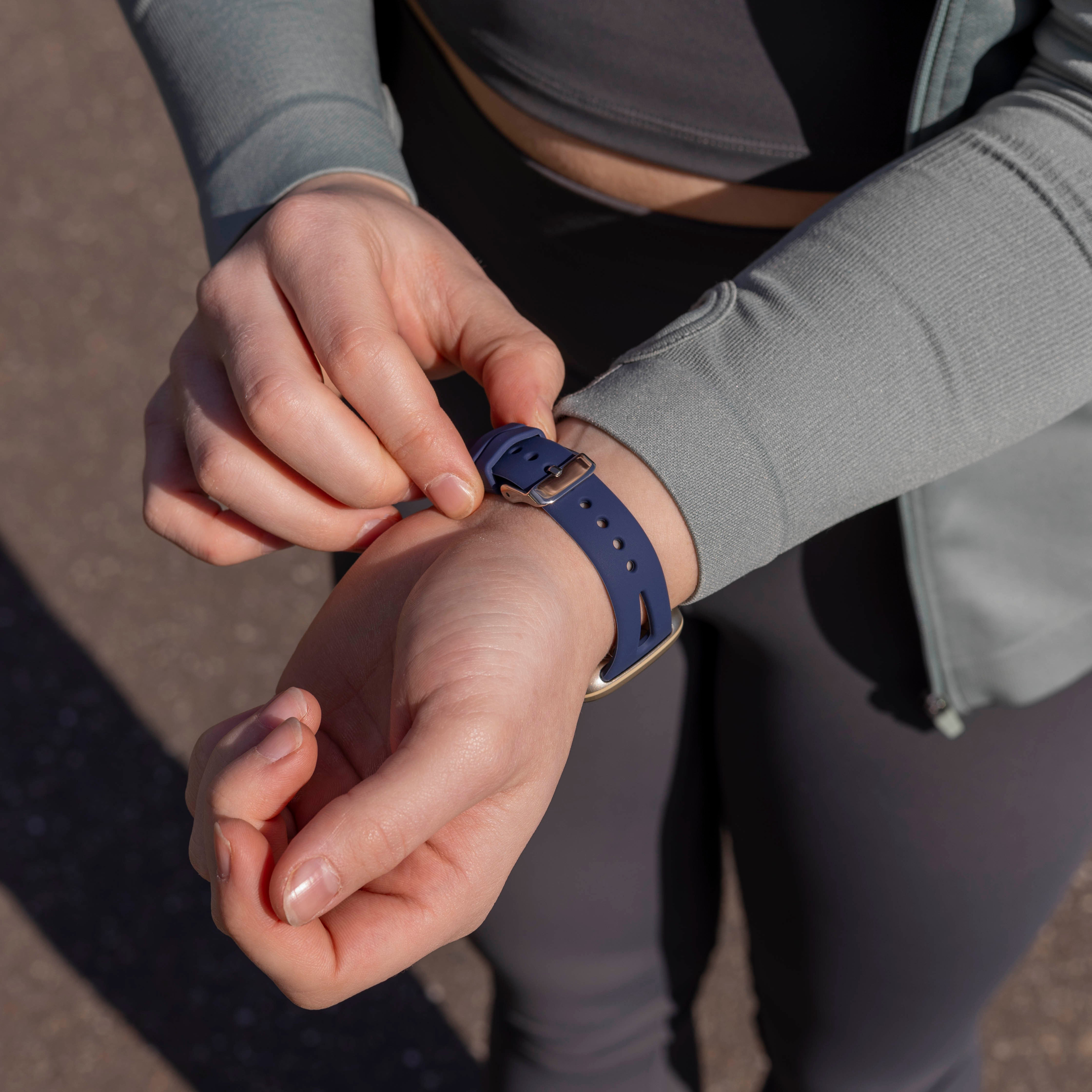Eine Person befestigt eine blaue Uhr an ihrem Handgelenk. Sie trägt eine graue Jacke und Leggings. Der Hintergrund ist unscharf.