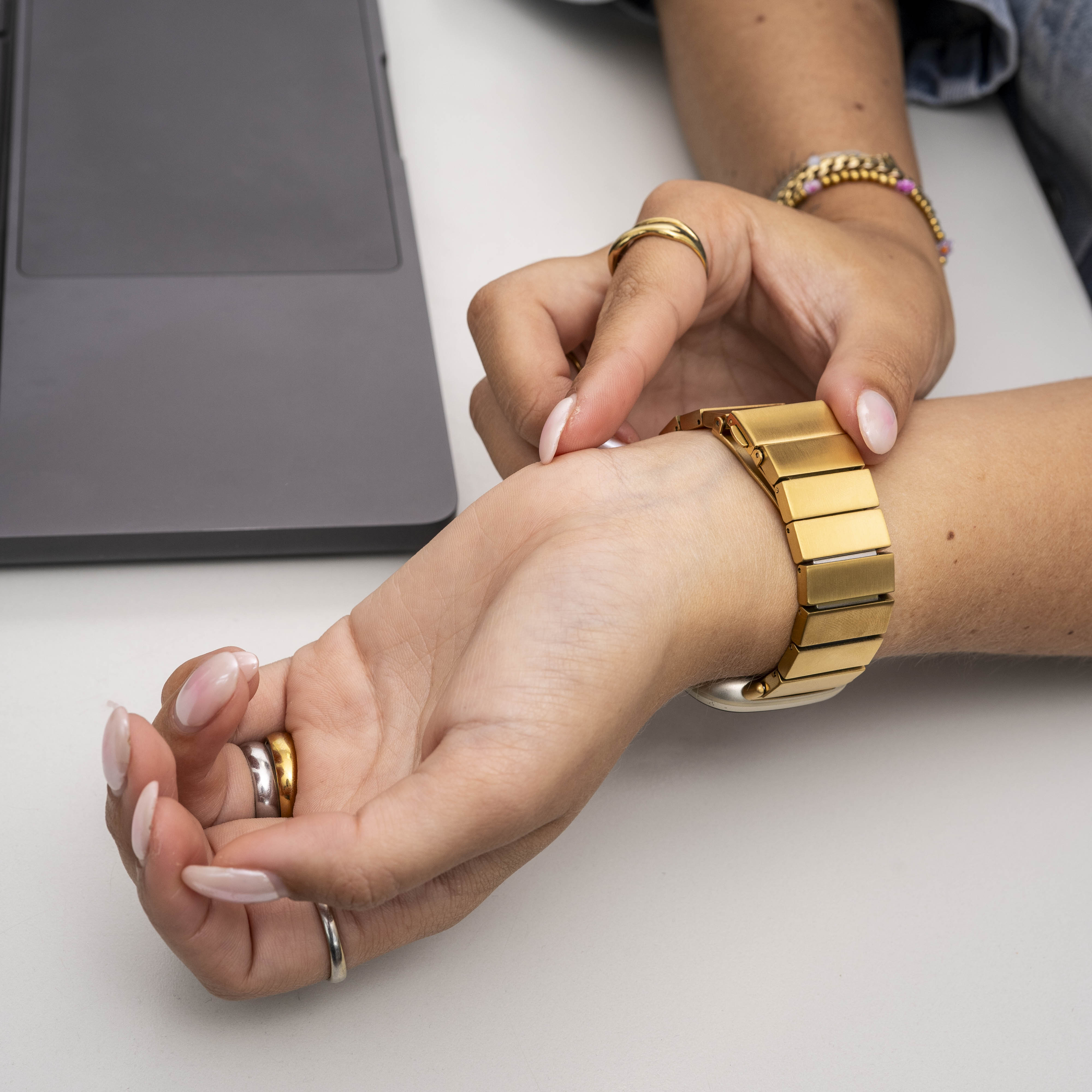 Eine Person justiert eine goldene Uhr. Hände mit Ringen sind sichtbar. Ein Laptop liegt auf der weißen Oberfläche.