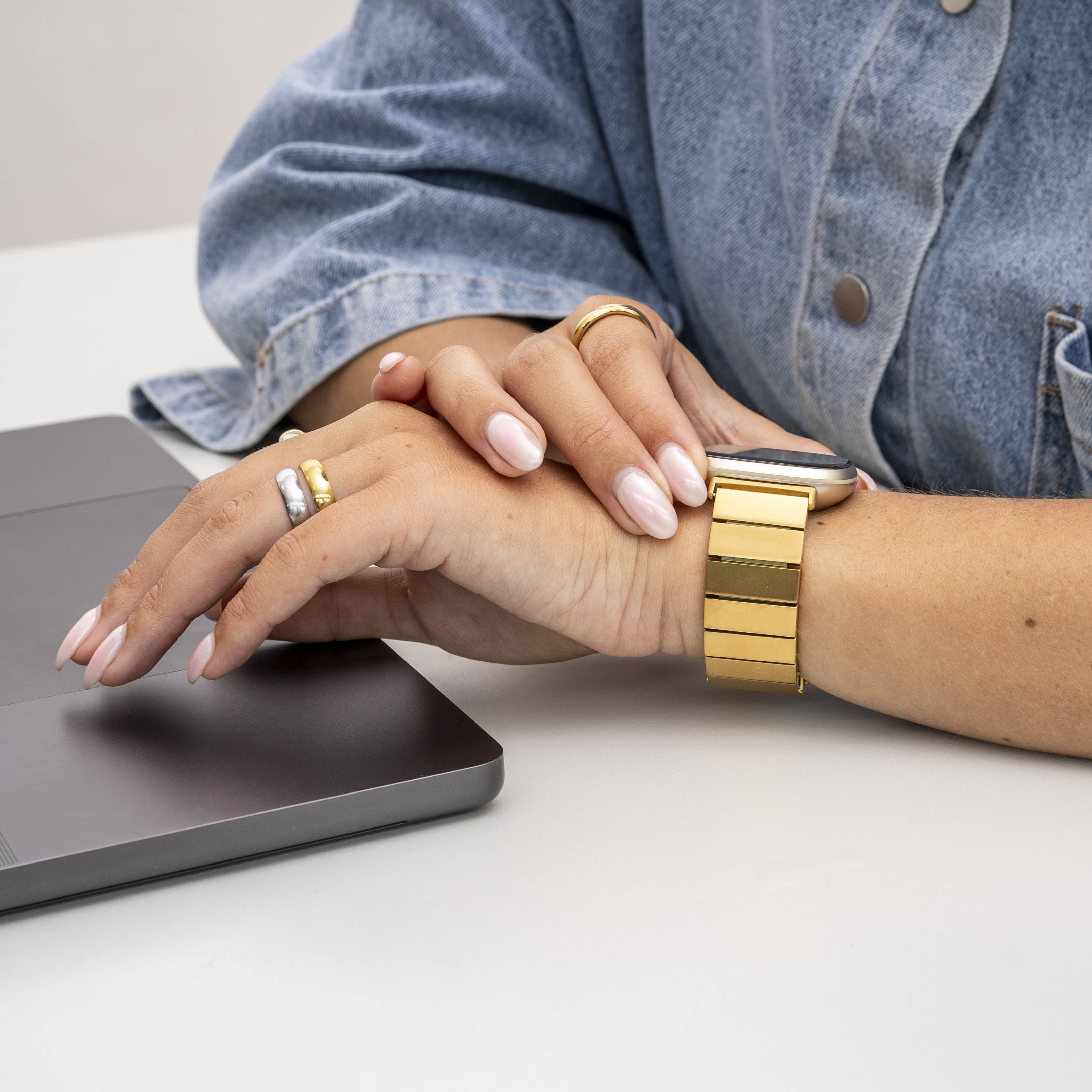 Hände mit Ringen ruhen auf einem Laptop. Eine goldene Uhr und eine Jeansjacke sind zu sehen. Nägel sind rosa lackiert.