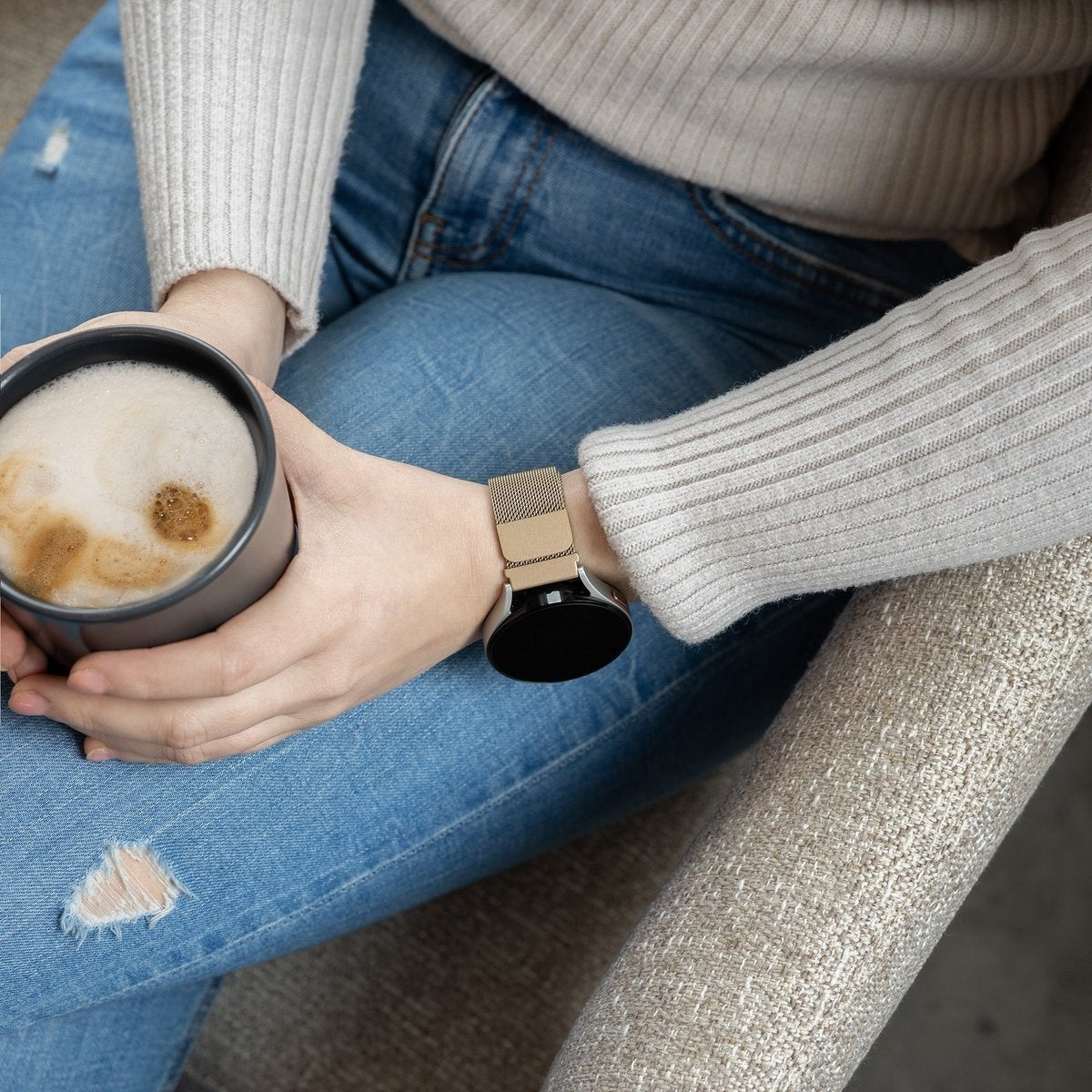 Person hält Kaffeetasse und trägt Smartwatch. Sie trägt einen beigen Pullover und blaue Jeans. Die Szene ist drinnen.