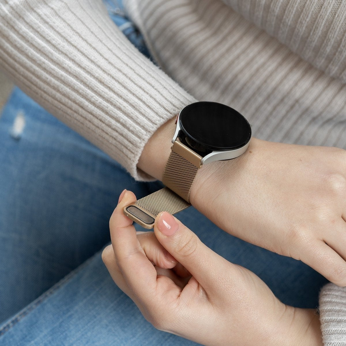 Eine Person trägt eine Smartwatch mit goldenem Armband. Die Person befestigt den Verschluss der Uhr. Der Hintergrund zeigt Jeans und einen Pullover.