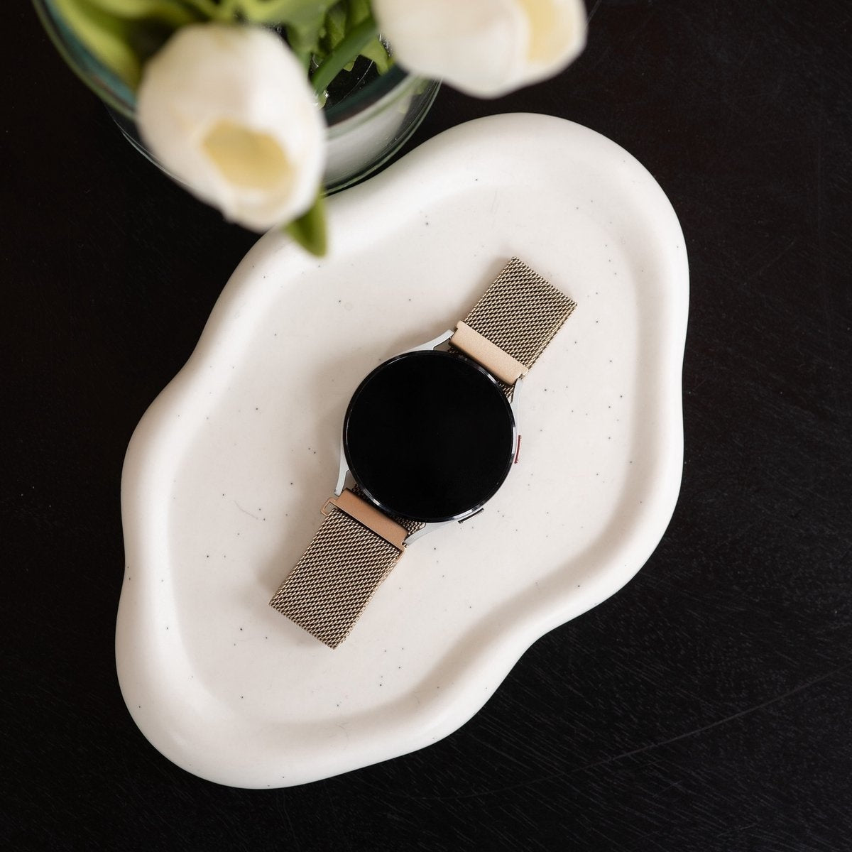 Eine Smartwatch mit goldenem Mesh-Band liegt auf einer weißen, organisch geformten Schale. Weiße Blumen in einem Glas oben links.