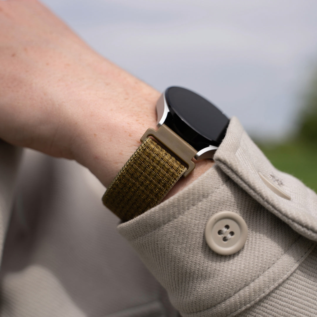 Das Handgelenk einer Person mit einer Smartwatch. Die Uhr hat einen schwarzen Bildschirm und ein olivgrünes Armband. Die Person trägt eine beige Jacke.