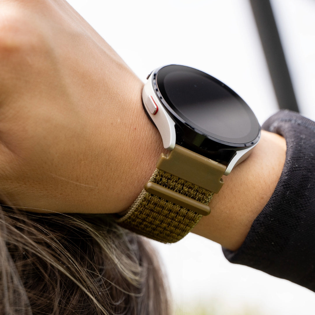 Das Handgelenk einer Person mit einer Smartwatch. Die Uhr hat einen weißen Rahmen, ein schwarzes Zifferblatt und ein olivgrünes Armband.