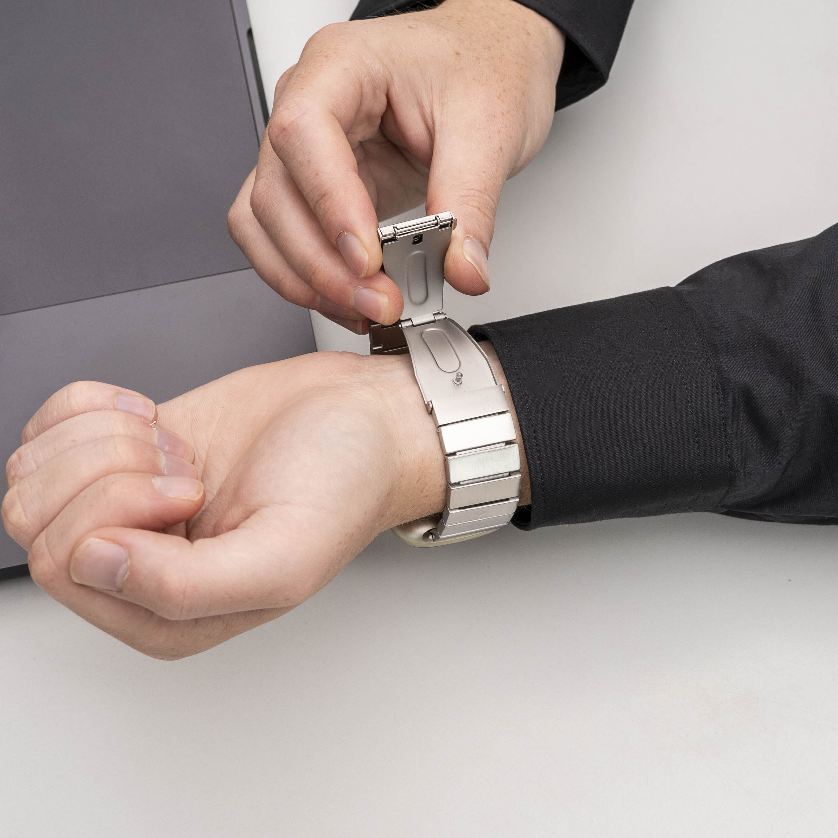 Eine Person befestigt eine silberne Armbanduhr an ihrem Handgelenk. Der Hintergrund zeigt einen Laptop und eine weiße Oberfläche.