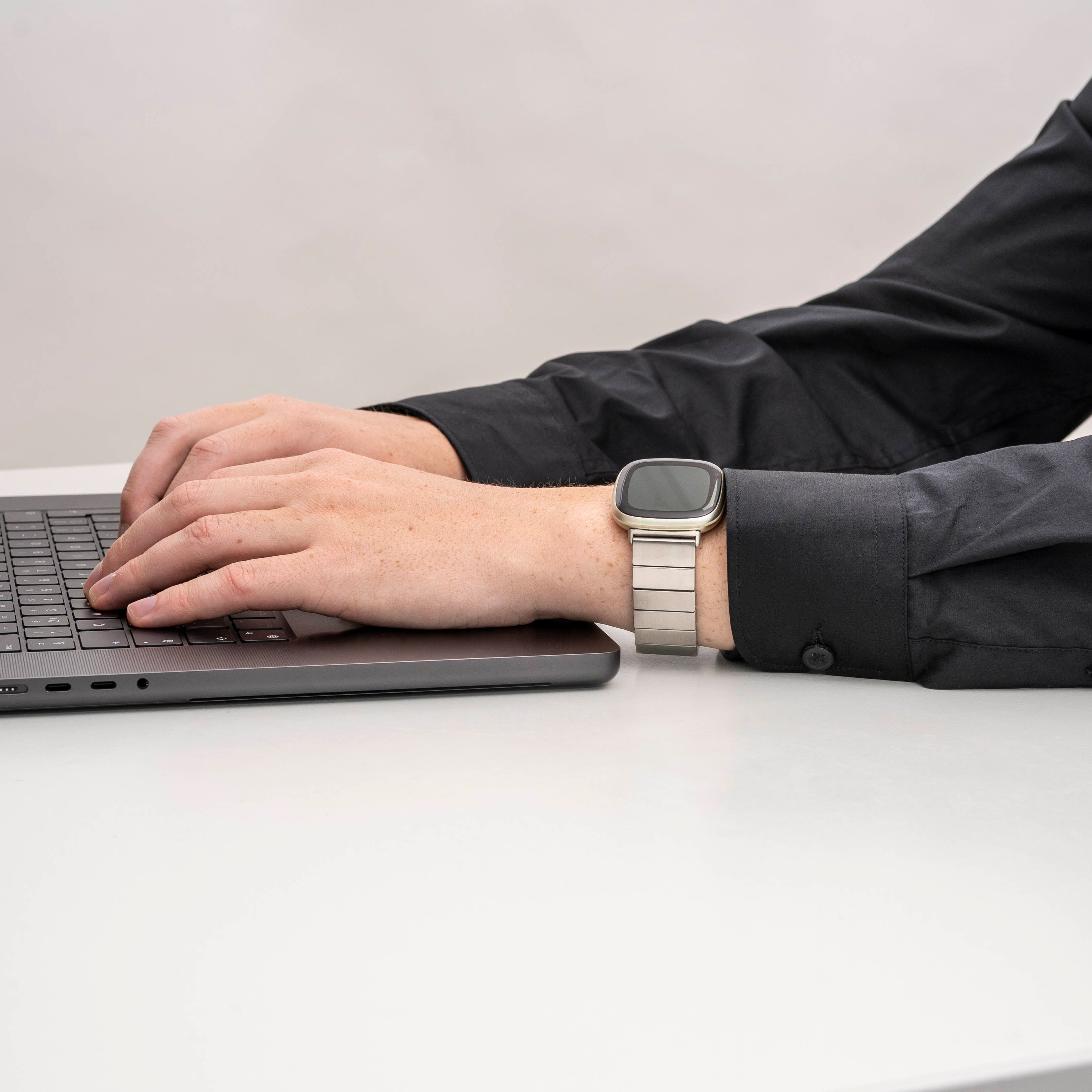 Eine Person tippt auf einem Laptop. Am Handgelenk eine Uhr. Schwarzes Hemd. Weiße Oberfläche.